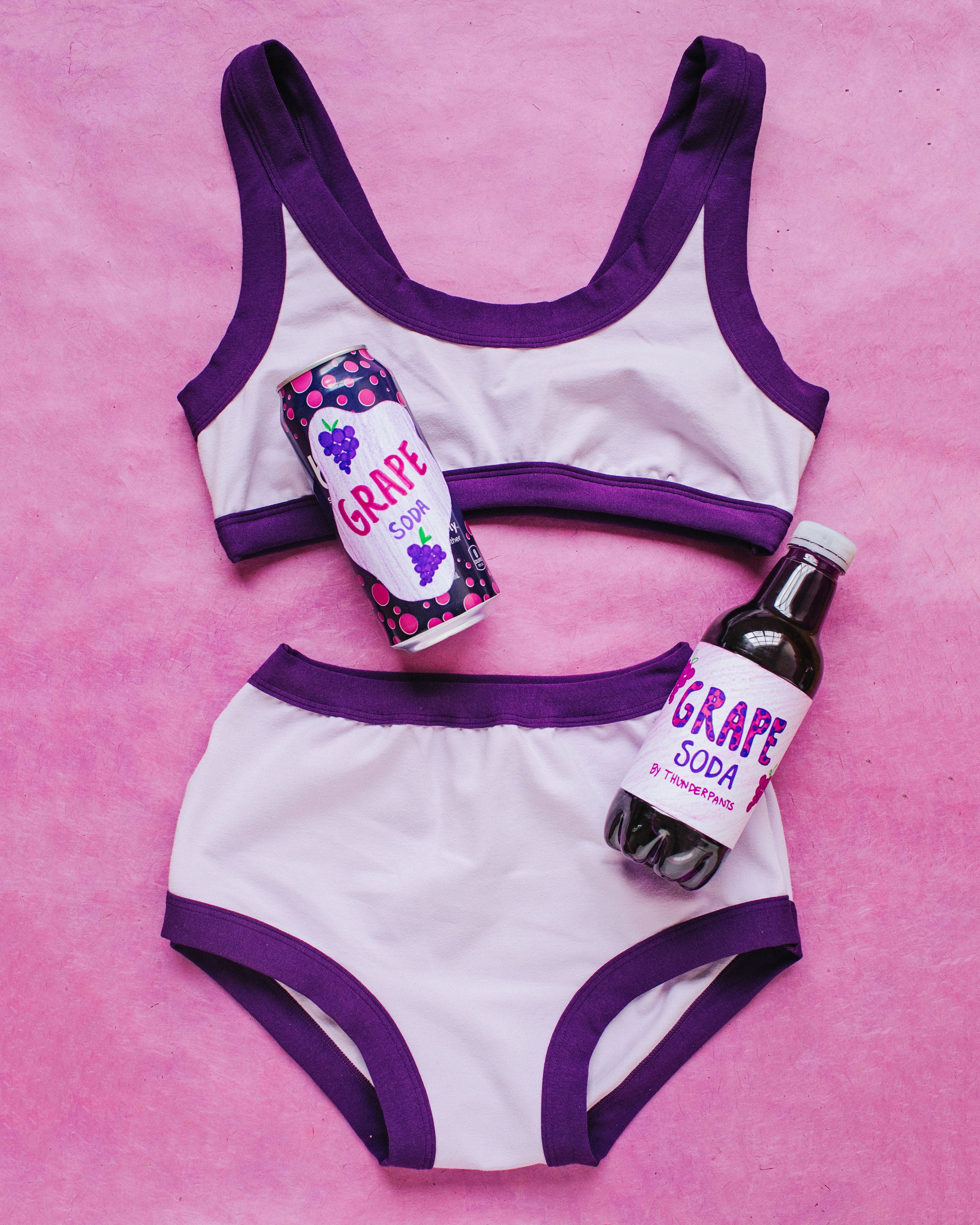 Flat lay with grape soda cans on purple surface of Grape Soda Bralette and Original style underwear set: light lavender with dark purple binding.