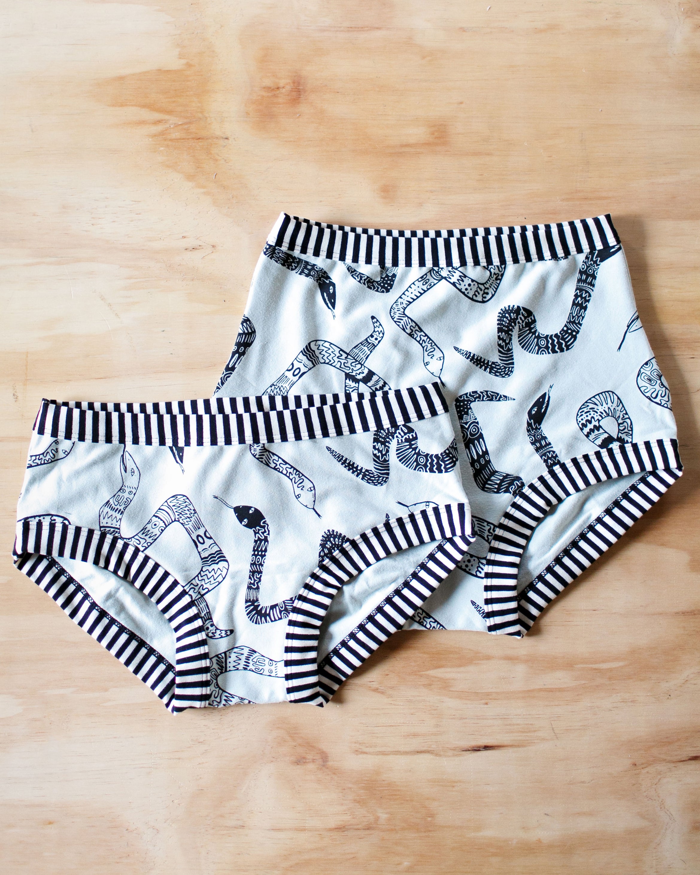 Flat lay of Thunderpants organic cotton Hipster style underwear and Sky Rise style underwear in Sketchy Snakes: sage color with black sketched snakes and black and white stripe binding.