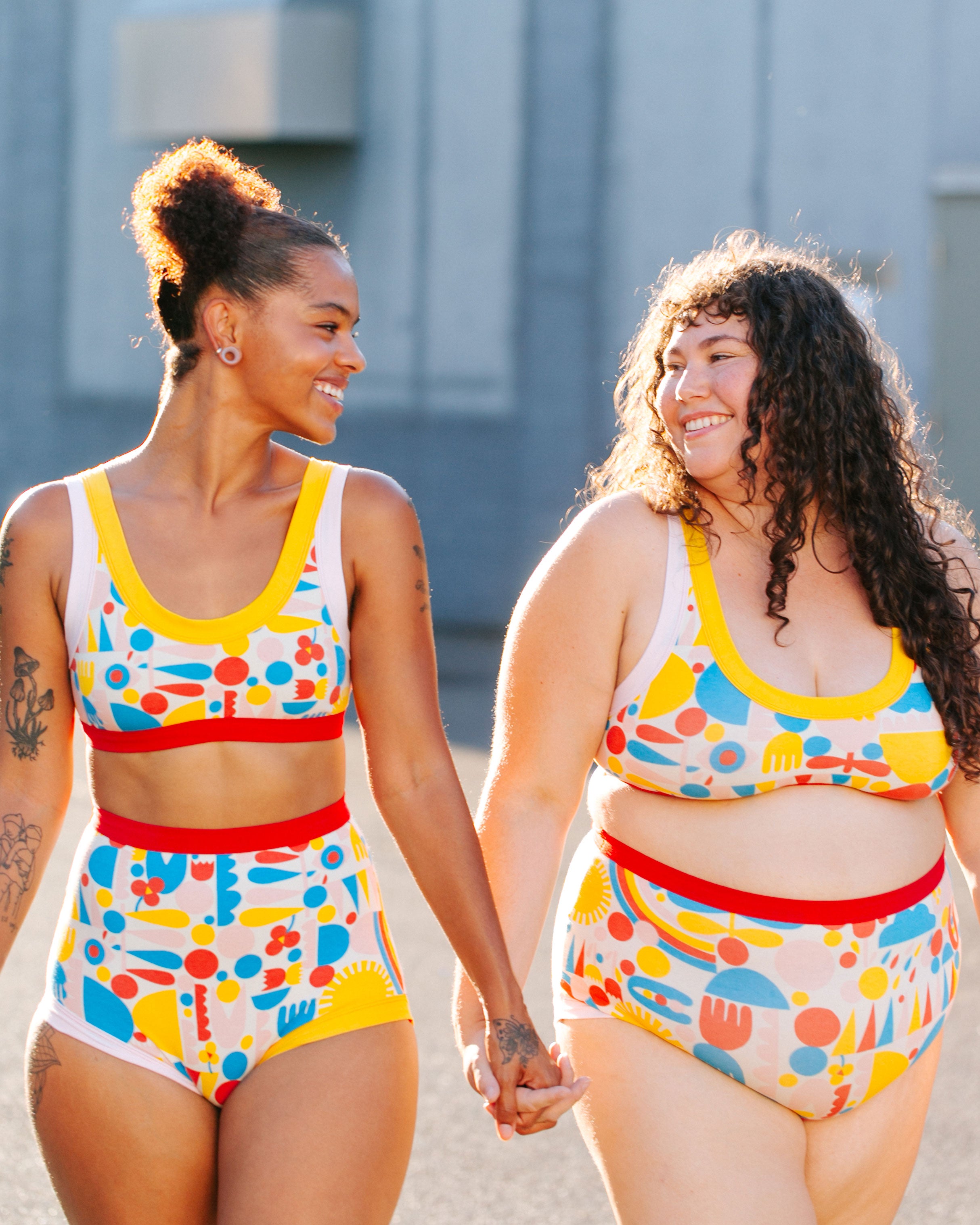 Models holding hands and smiling in the sun wearing sets of Bralettes, Sky Rise and Original style underwear in Balance by Lisa Congdon: geometric shapes in red, blue, yellow, and pink colors.