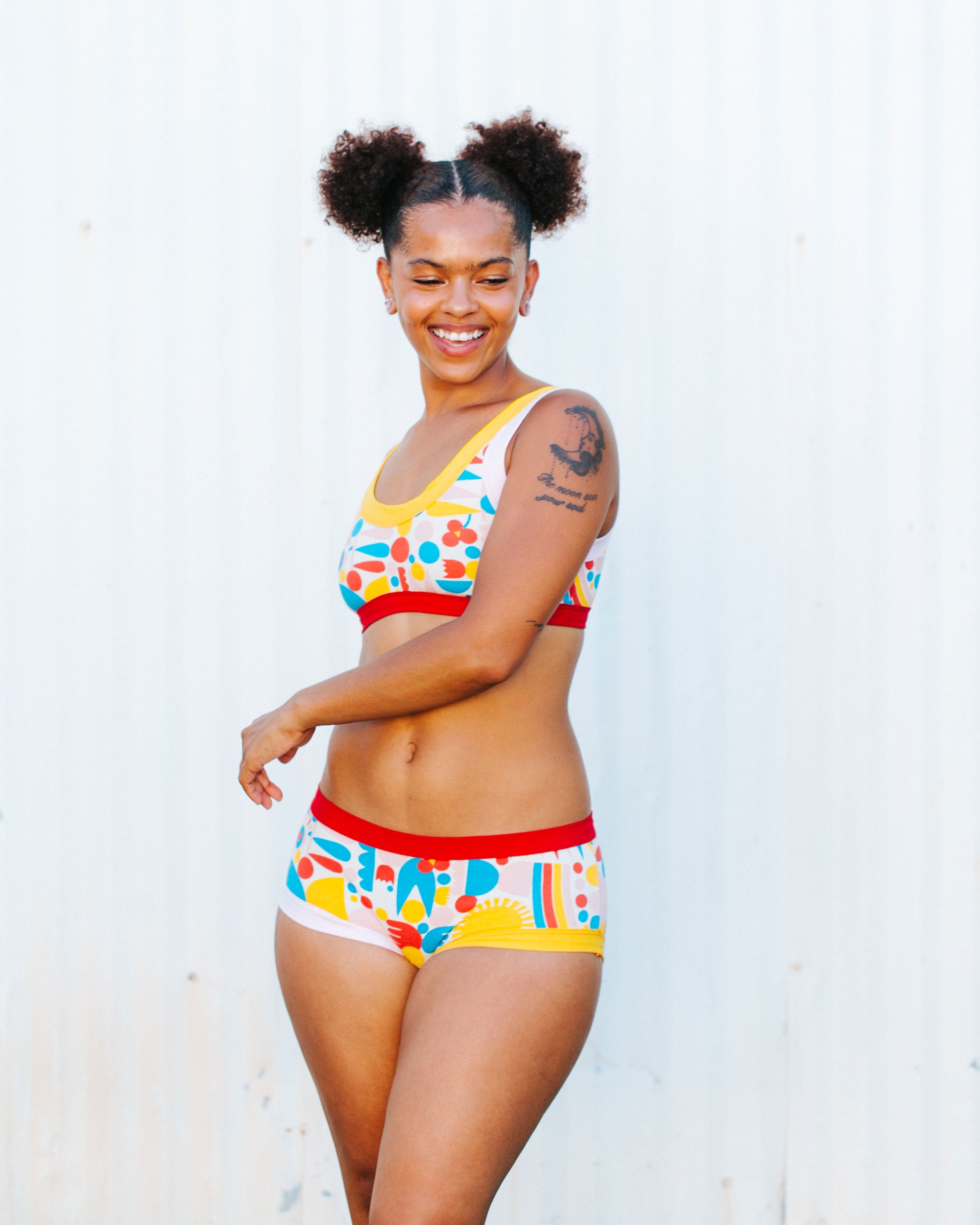 Model smiling in front of a white wall wearing a set of Hipster style underwear and Bralette in Balance by Lisa Congdon print: geometric shapes in red, pink, yellow, and blue colors.