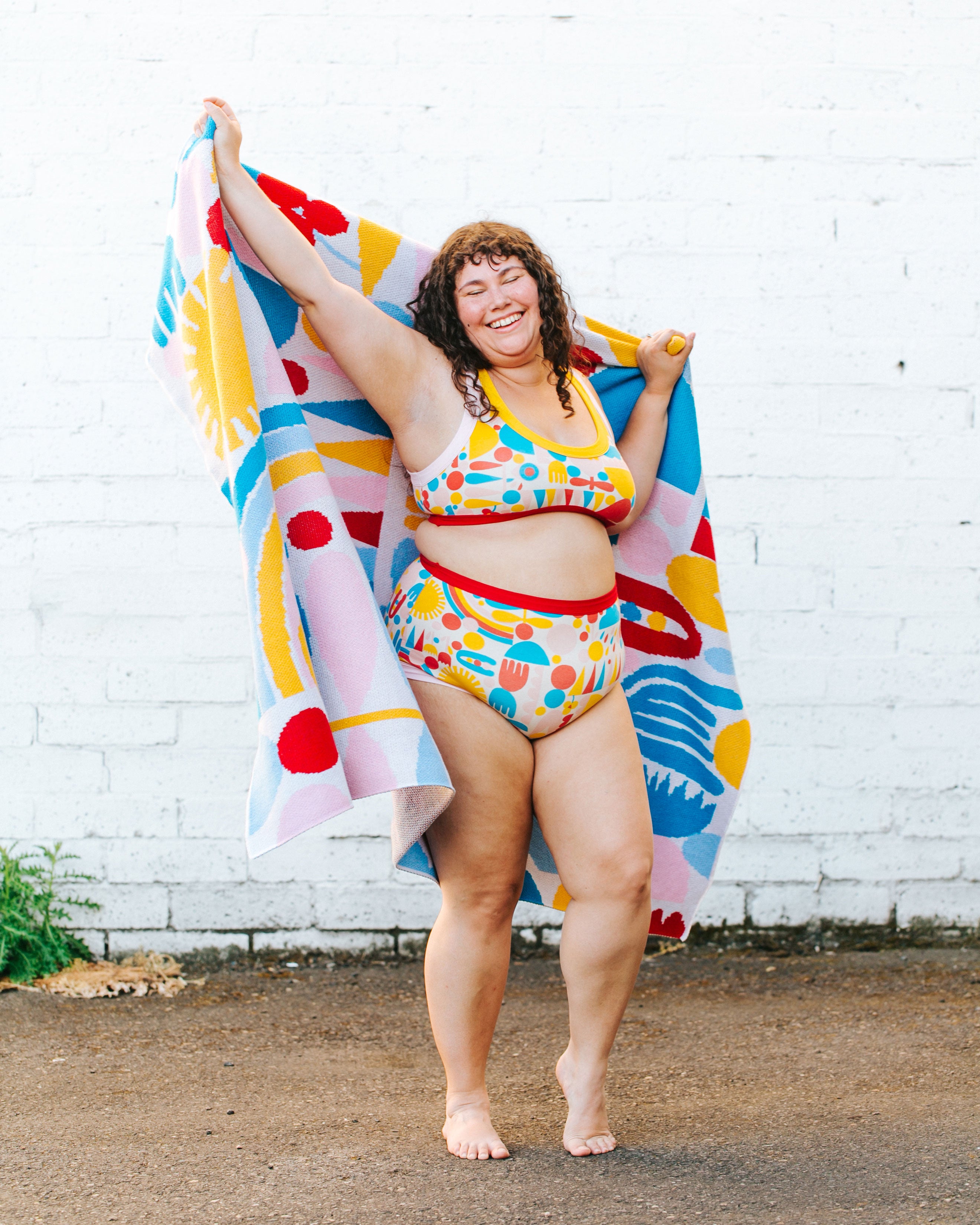 Model smiling and dancing in a set of Original style underwear and Bralette in Balance by Lisa Congdon print: geometric shapes in red, pink, yellow, and blue colors, with a similar blanket around her shoulders.