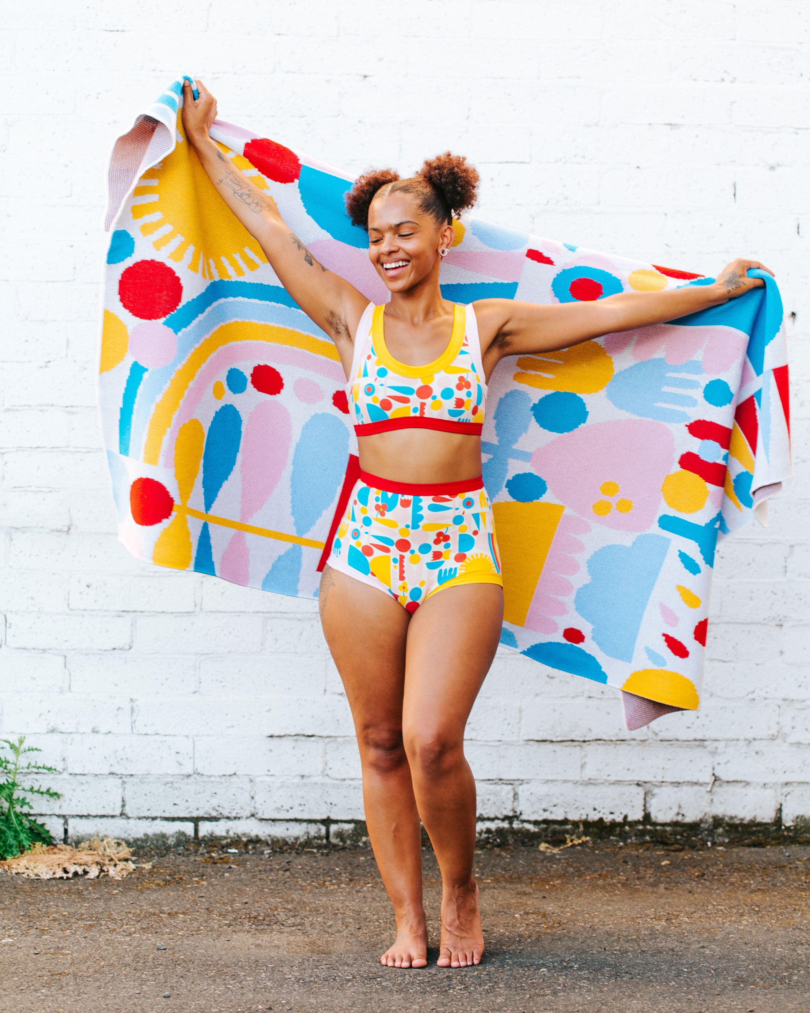 Model smiling while holding a blanket wearing a set of Sky Rise style underwear and Bralette in Balance by Lisa Congdon print: geometric shapes in pink, red, blue, and yellow colors.