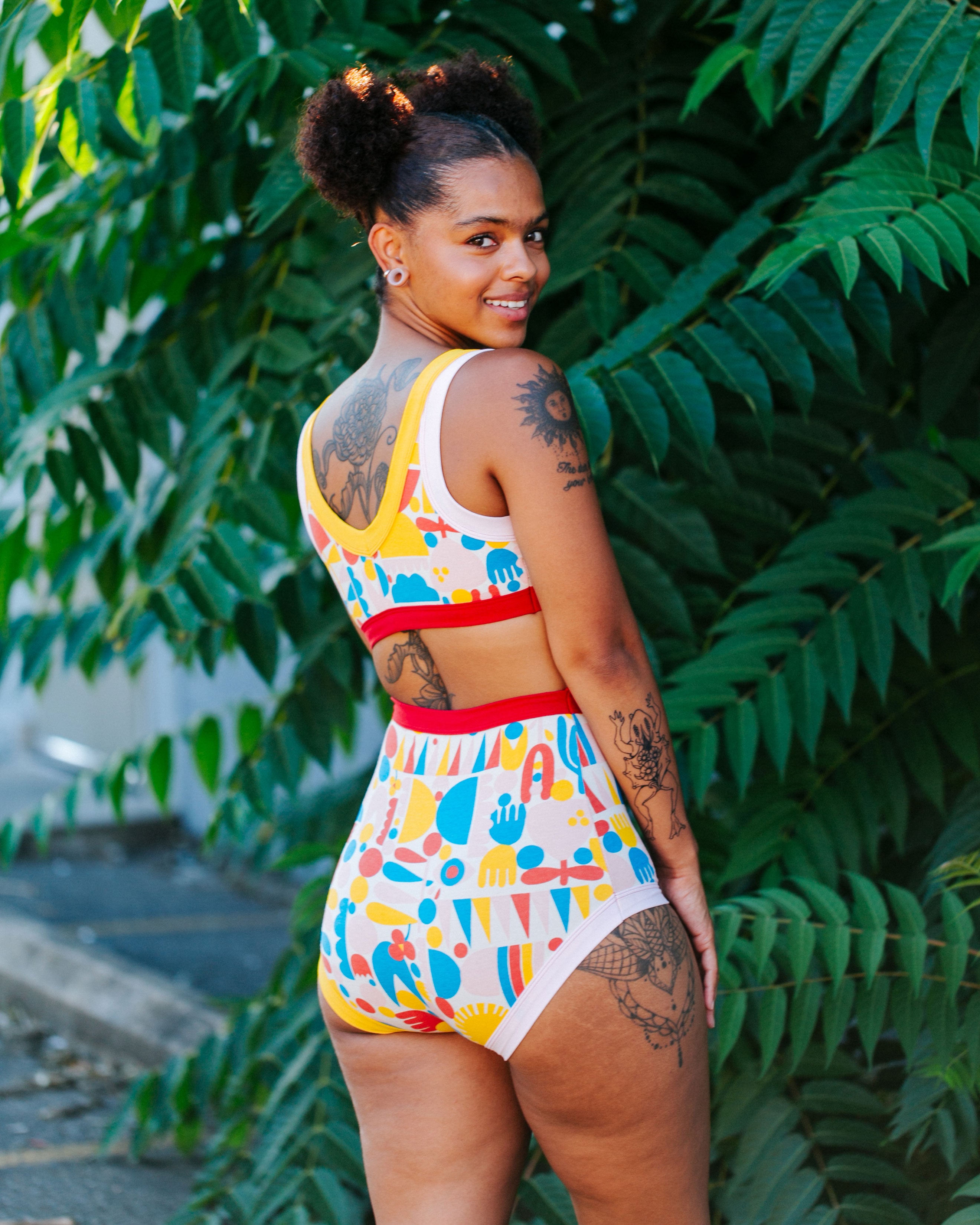 Model with her back to us in front of tropical plants wearing a set of Sky Rise style underwear and Bralette in Balance by Lisa Congdon print: geometric shapes in pink, red, yellow, and blue colors. 