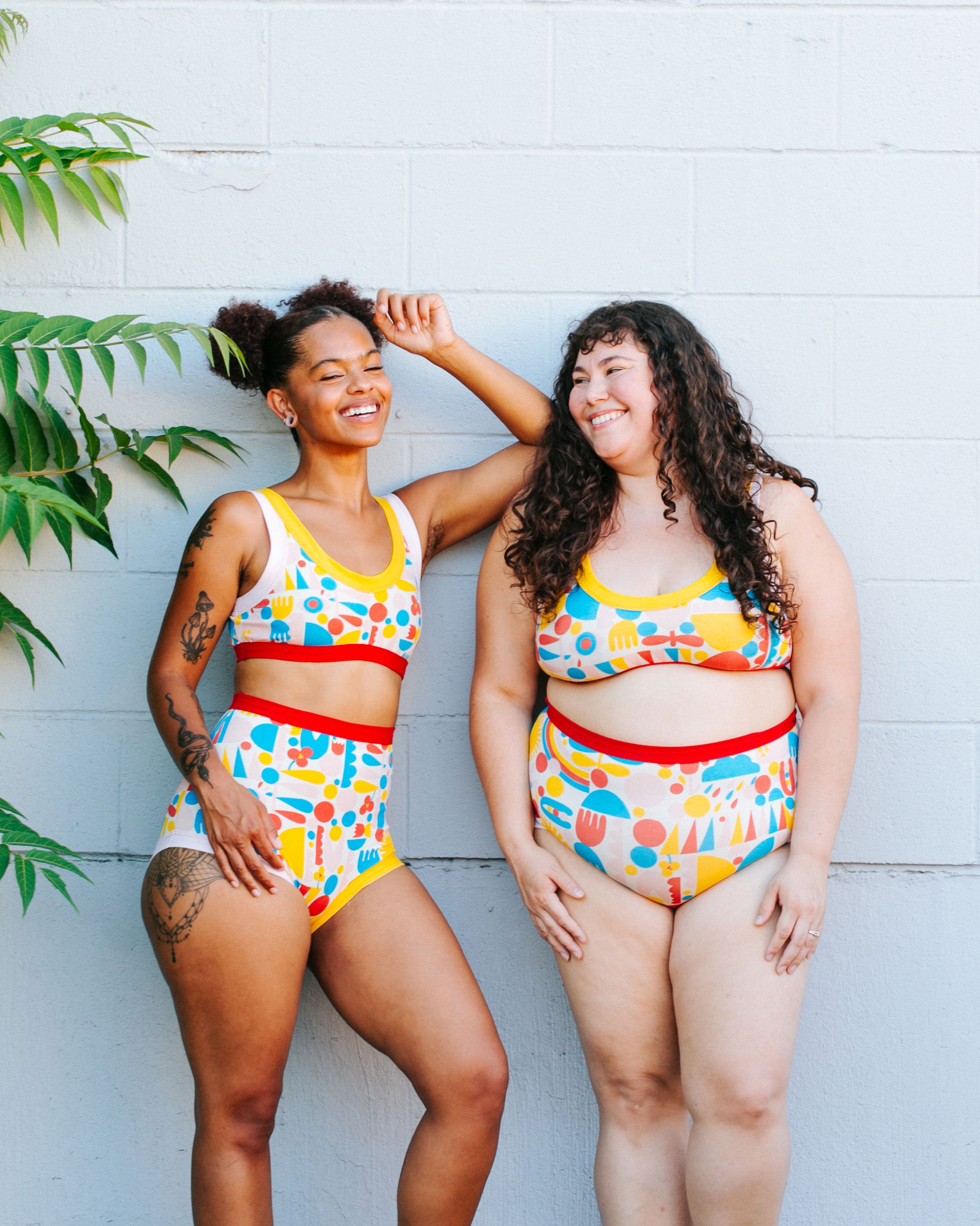 Two models laughing together against a white brick wall wearing sets of Sky Rise style and Original style underwear with Bralettes in Balance by Lisa Congdon print: geometric shapes in red, pink, yellow, and blue colors.