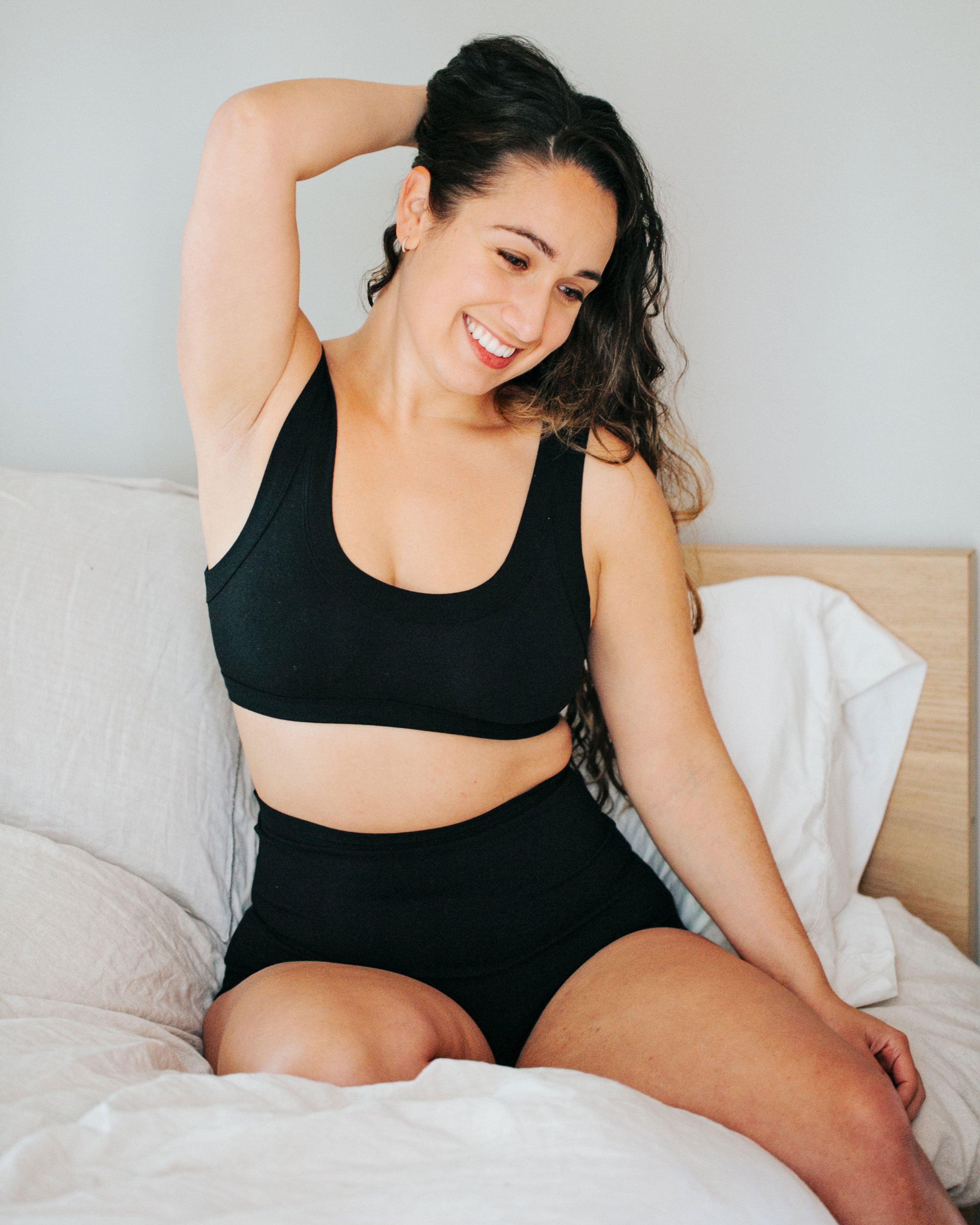 Model sitting on a bed smiling wearing Plain Black Sky Rise underwear and Bralette set.