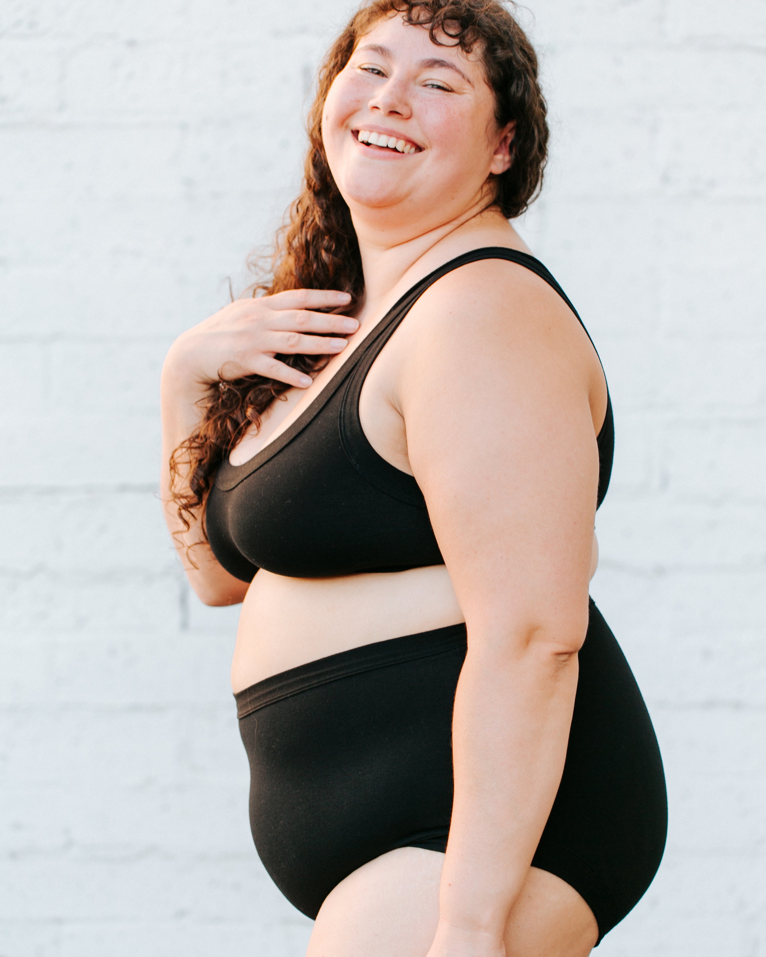 Plus sized model smiling into the camera wearing Original style plain black underwear and Bralette set.