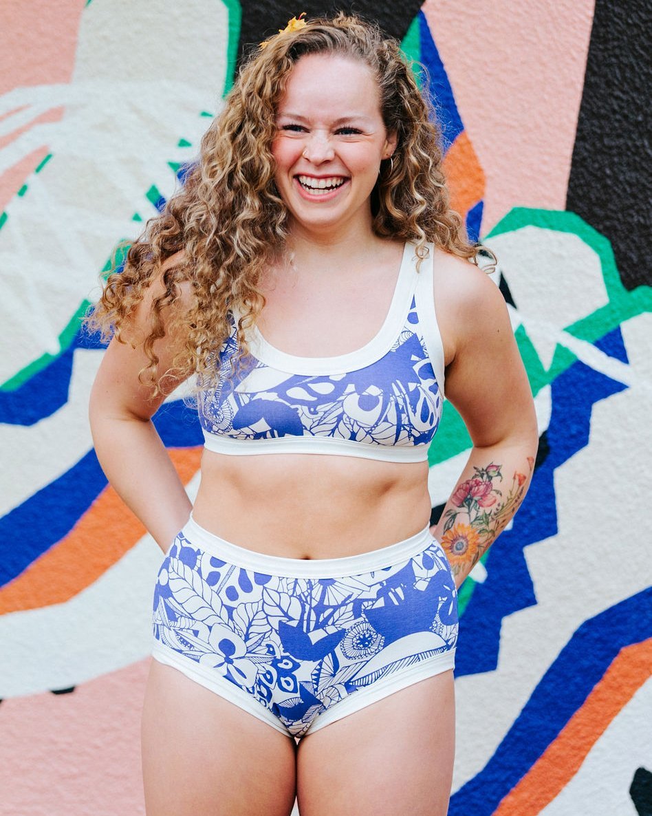 Laughing woman wearing Thunderpants organic cotton Original style underwear and Bralette in a blue jungle and floral print with a colorful background.