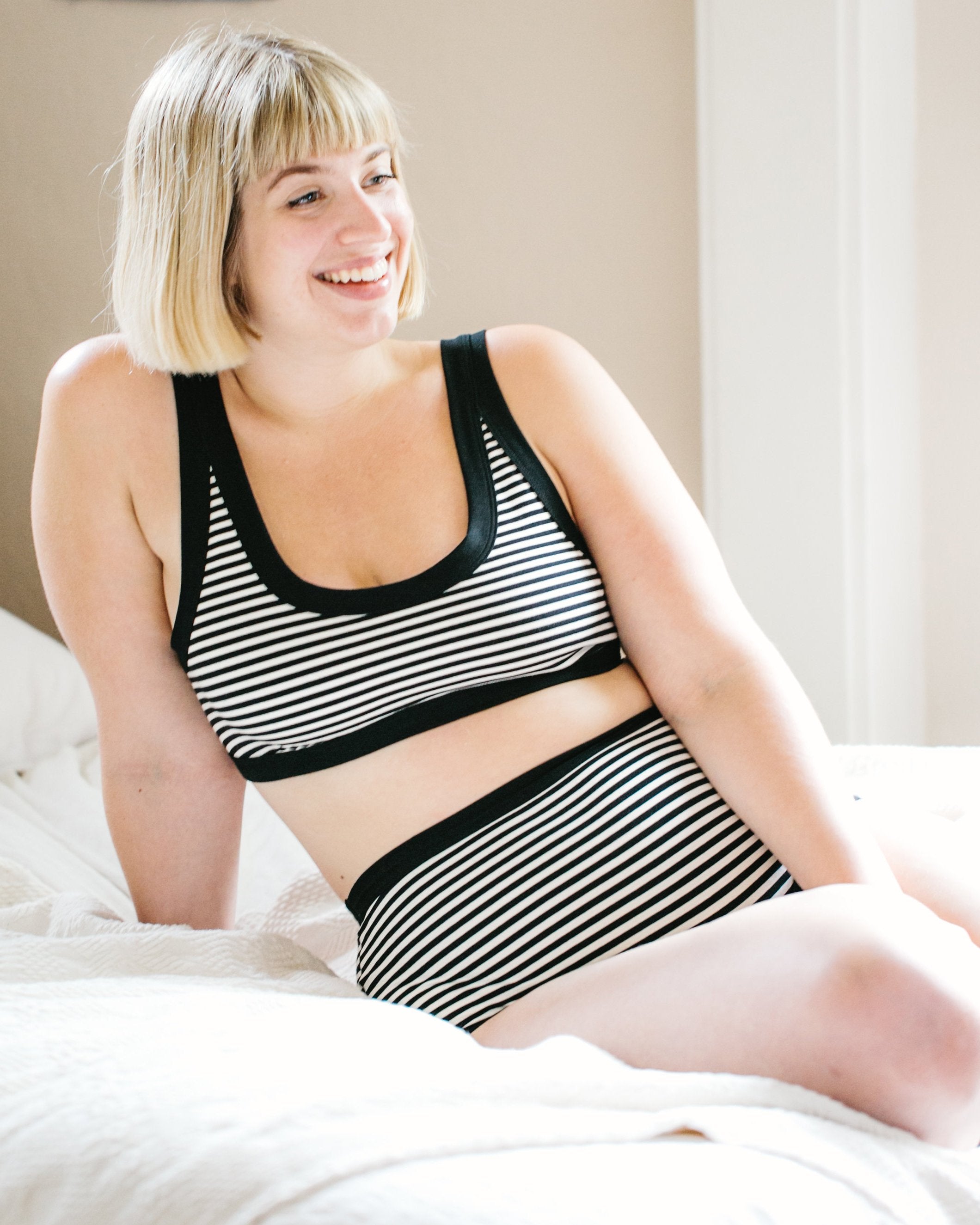 Model happily lounging on a bed wearing Thunderpants organic cotton Bralette and Original style underwear in black and white stripes.