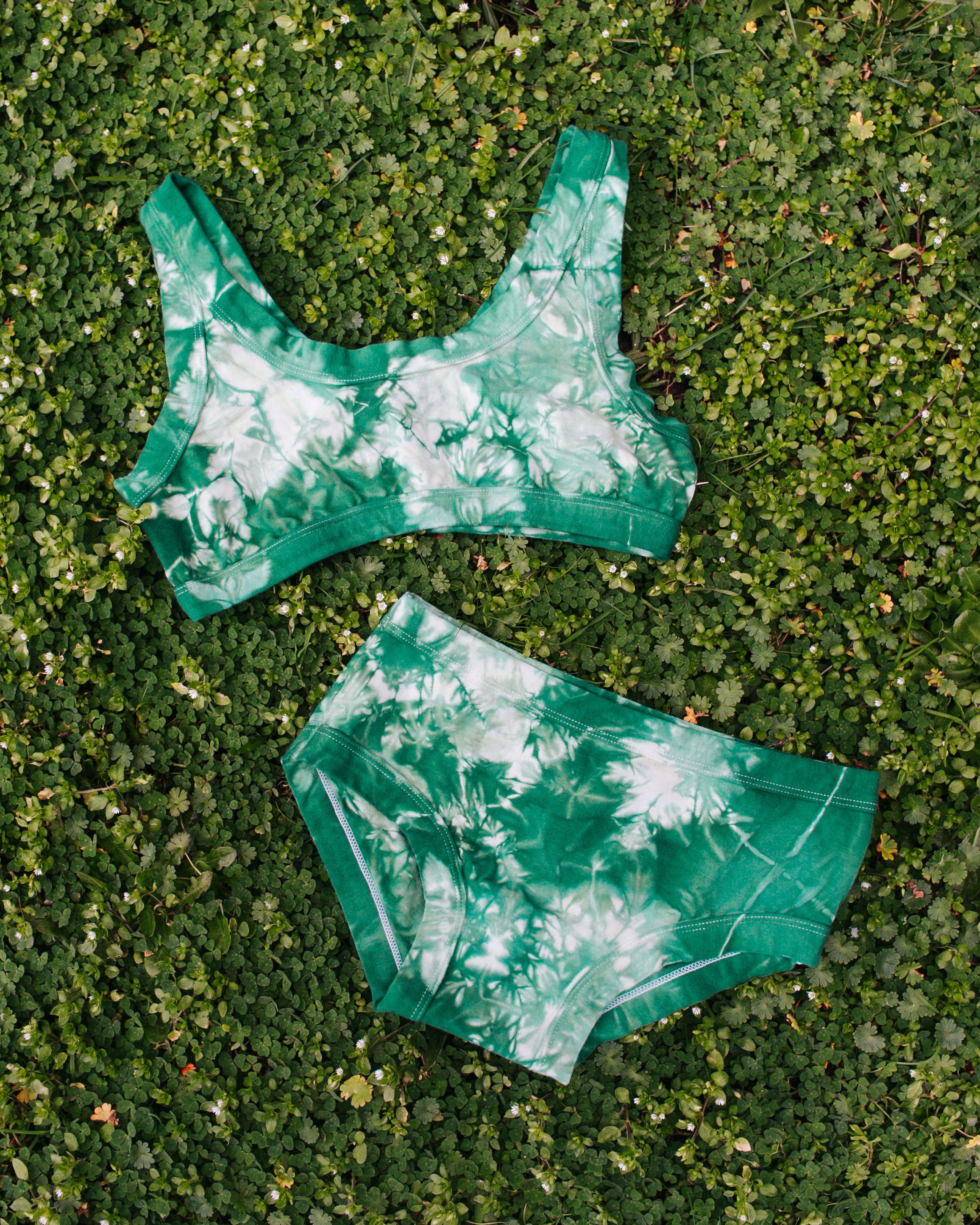 Flat lay on bright green clover grass of Thunderpants organic cotton Bralette and Hipster style underwear in hand dye Clover Green scrunch dye.
