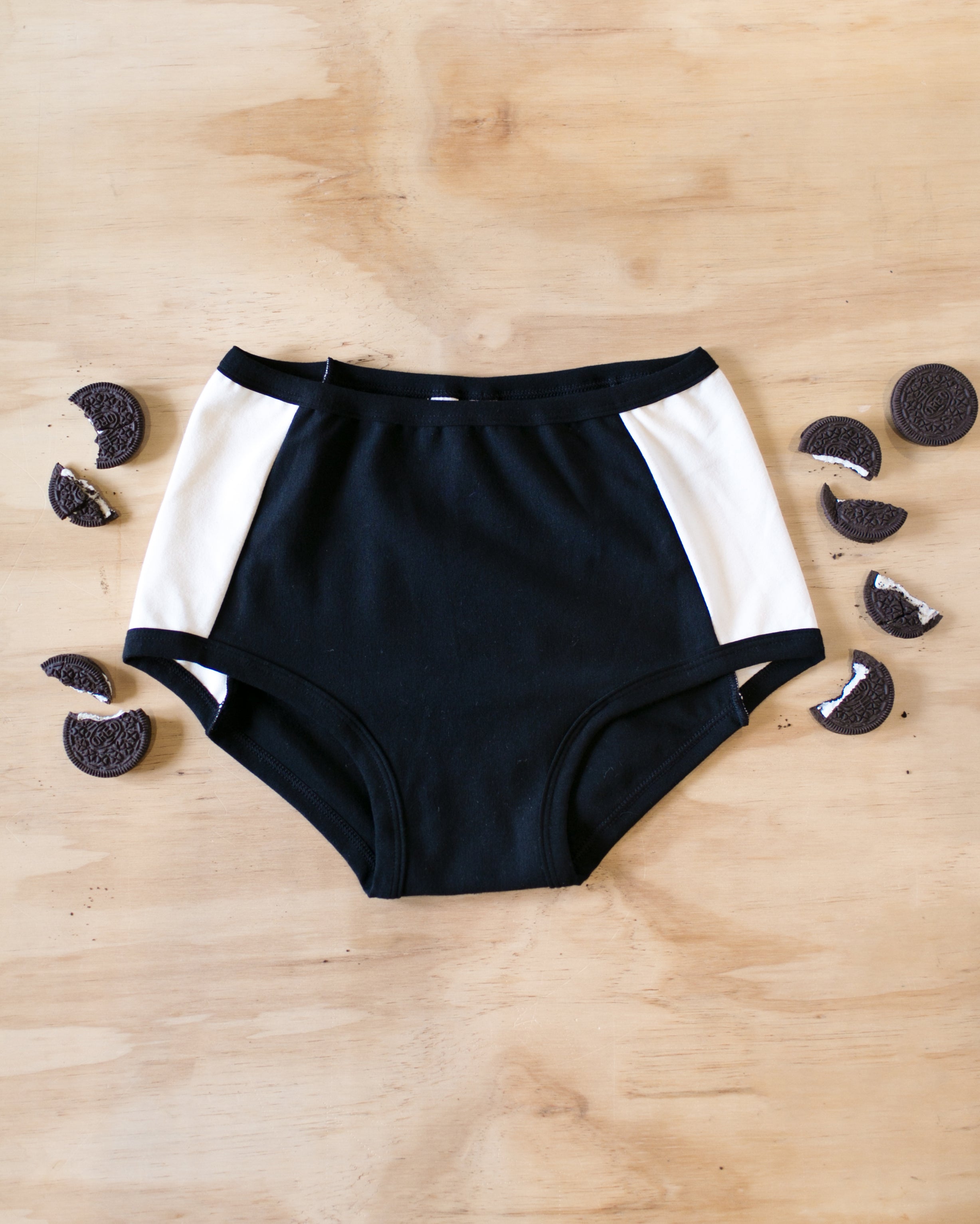 Flat lay on wood of our Cookies and Cream Panel Pants in the Original style with Oreo cookies surrounding it: black panel in the center and vanilla panels on both sides with black binding.