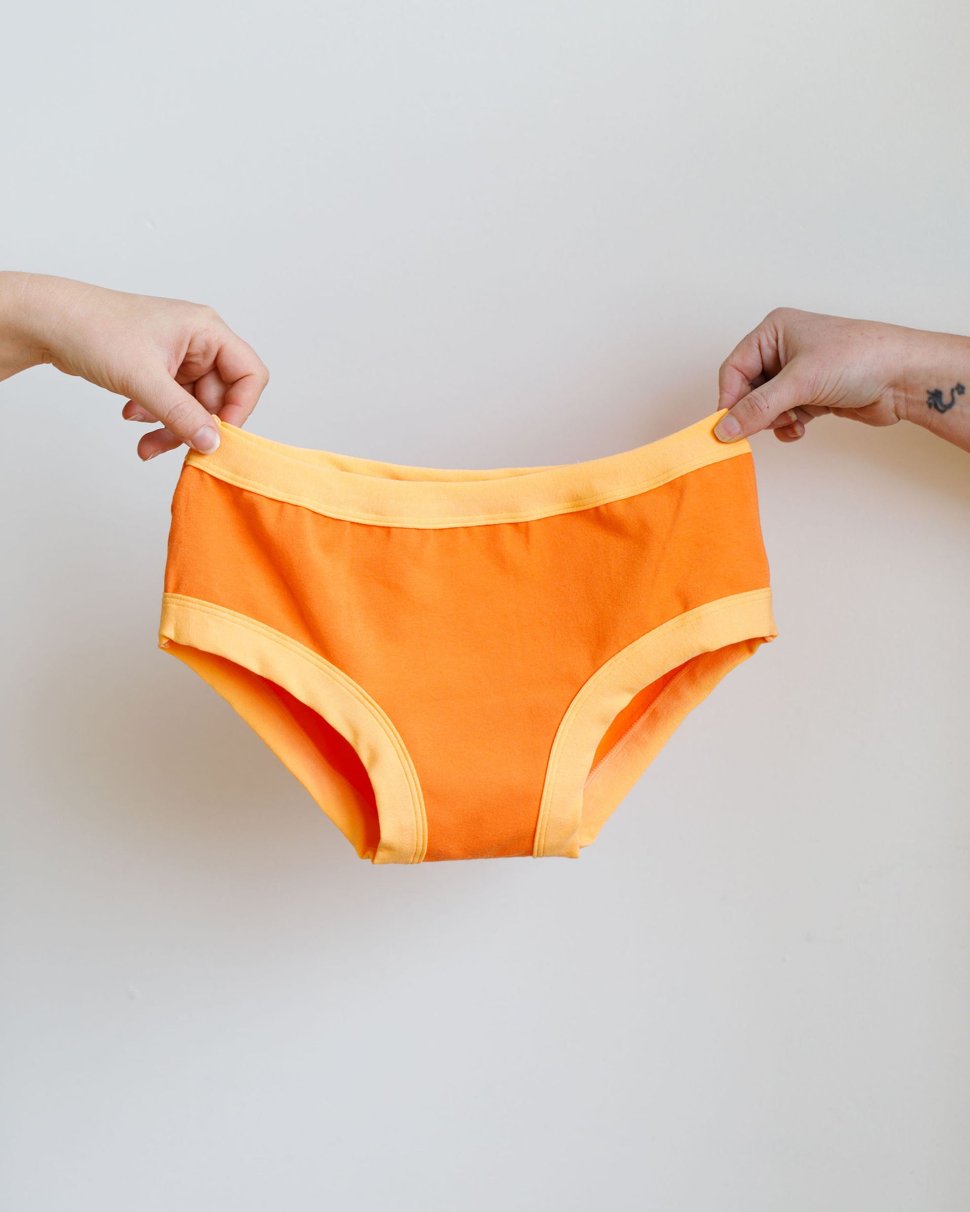 Two hands holding Thunderpants Hipster style underwear in Creamsicle: dark orange with light orange binding.