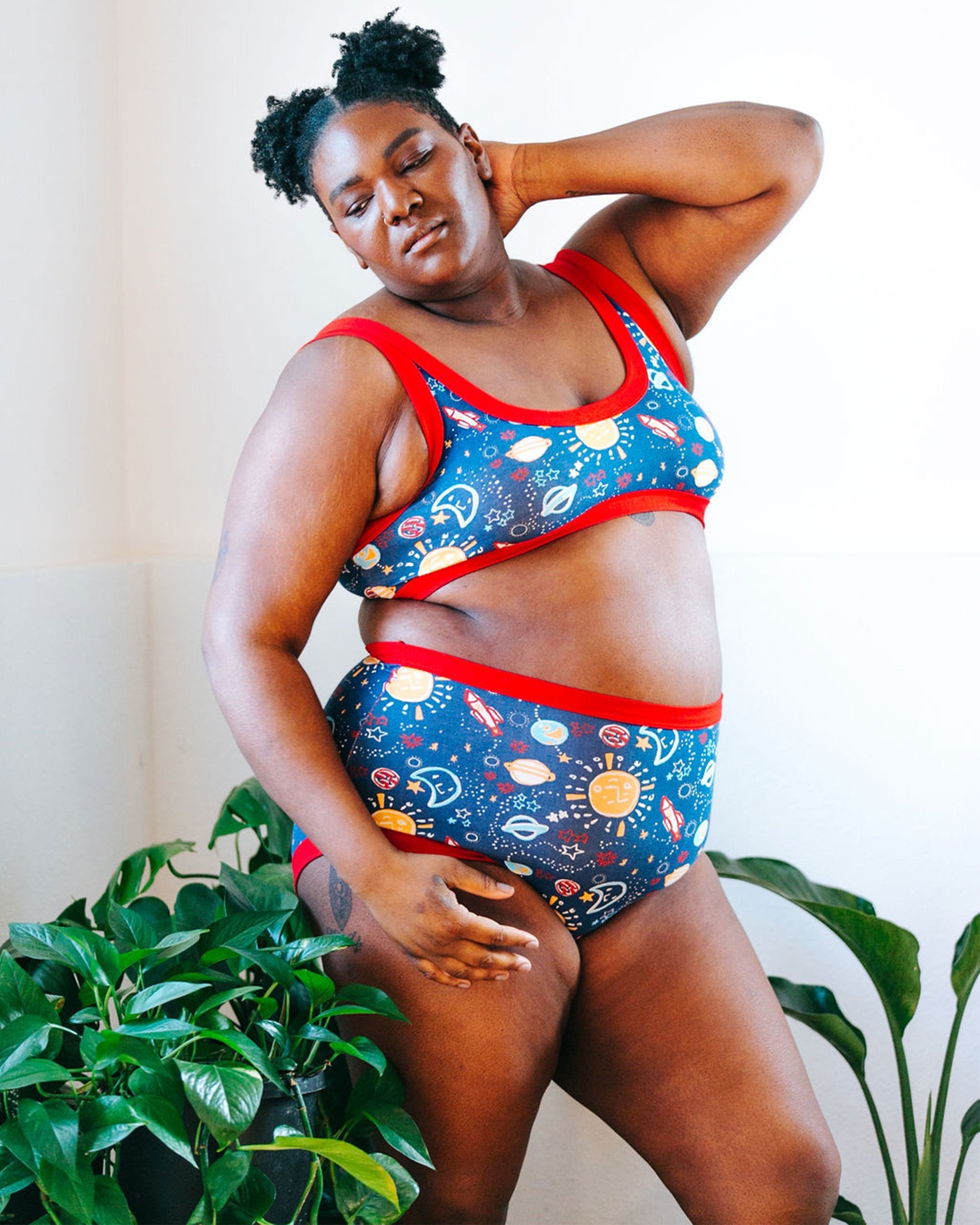 Plus-sized model wearing Thunderpants organic cotton Original style underwear and Bralette in a sun, planets, stars, and universe print with plants around her.