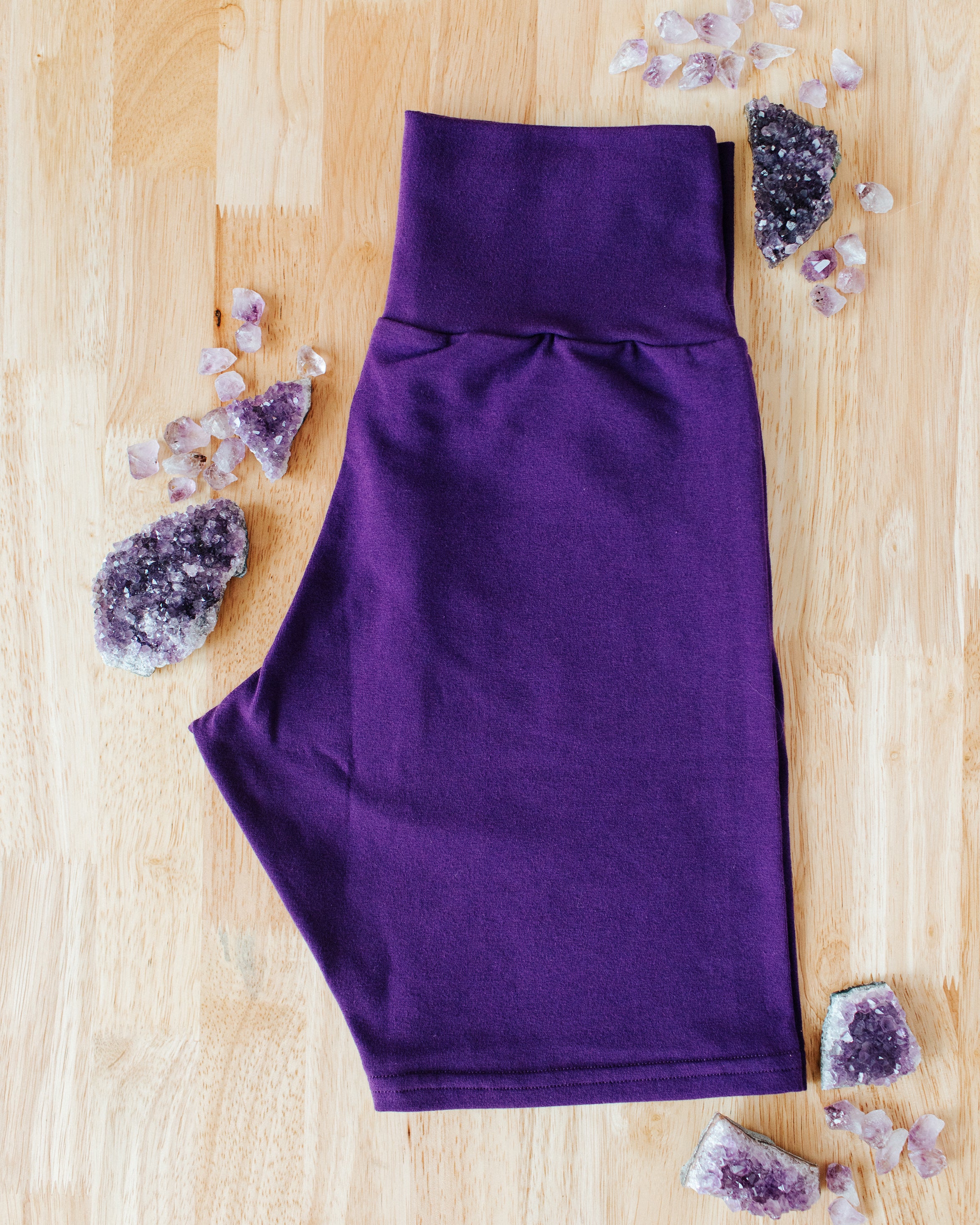 Flat lay of folded purple Deep Amethyst Bike Shorts on a wood surface with big and small amethyst stones surrounding it.