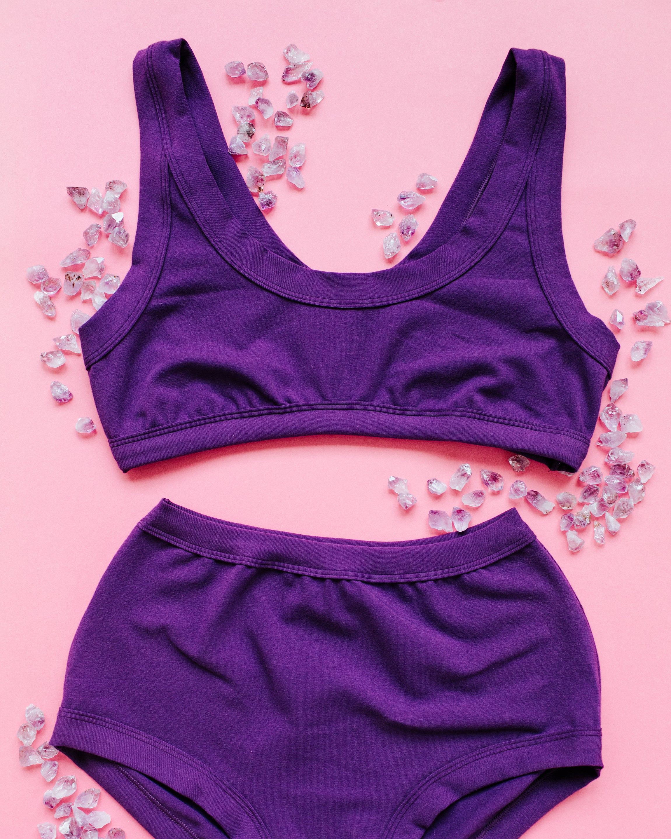 Flat lay of purple Deep Amethyst Bralette and Original style underwear on a pink backdrop with small amethyst stones surrounding it.