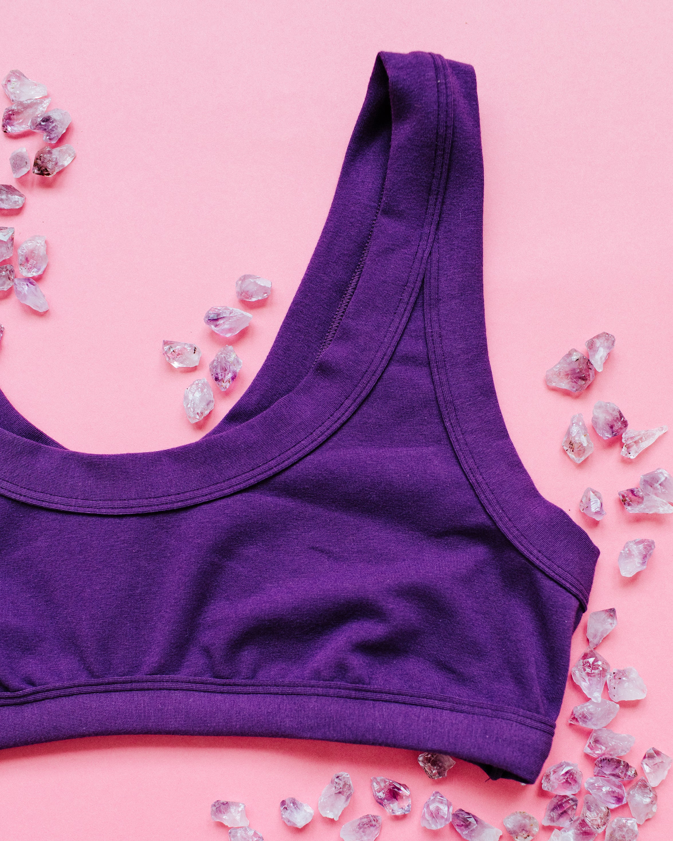 Flat lay of purple Deep Amethyst Bralette on a pink background with small amethyst stones surrounding it.