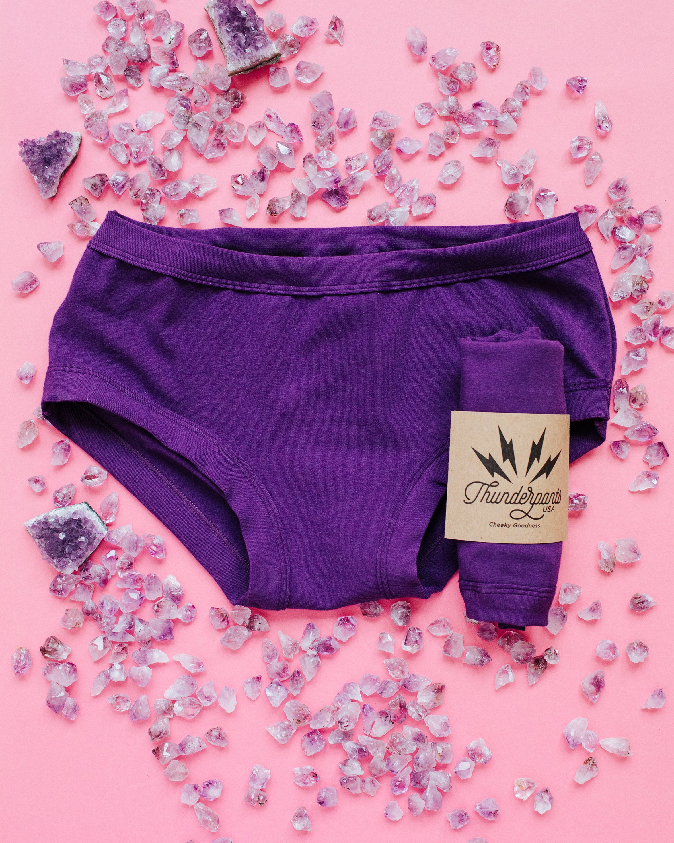 Flat lay of purple Deep Amethyst Hipster style underwear and packaged underwear on a pink surface with lots of small and large amethyst stones surrounding.