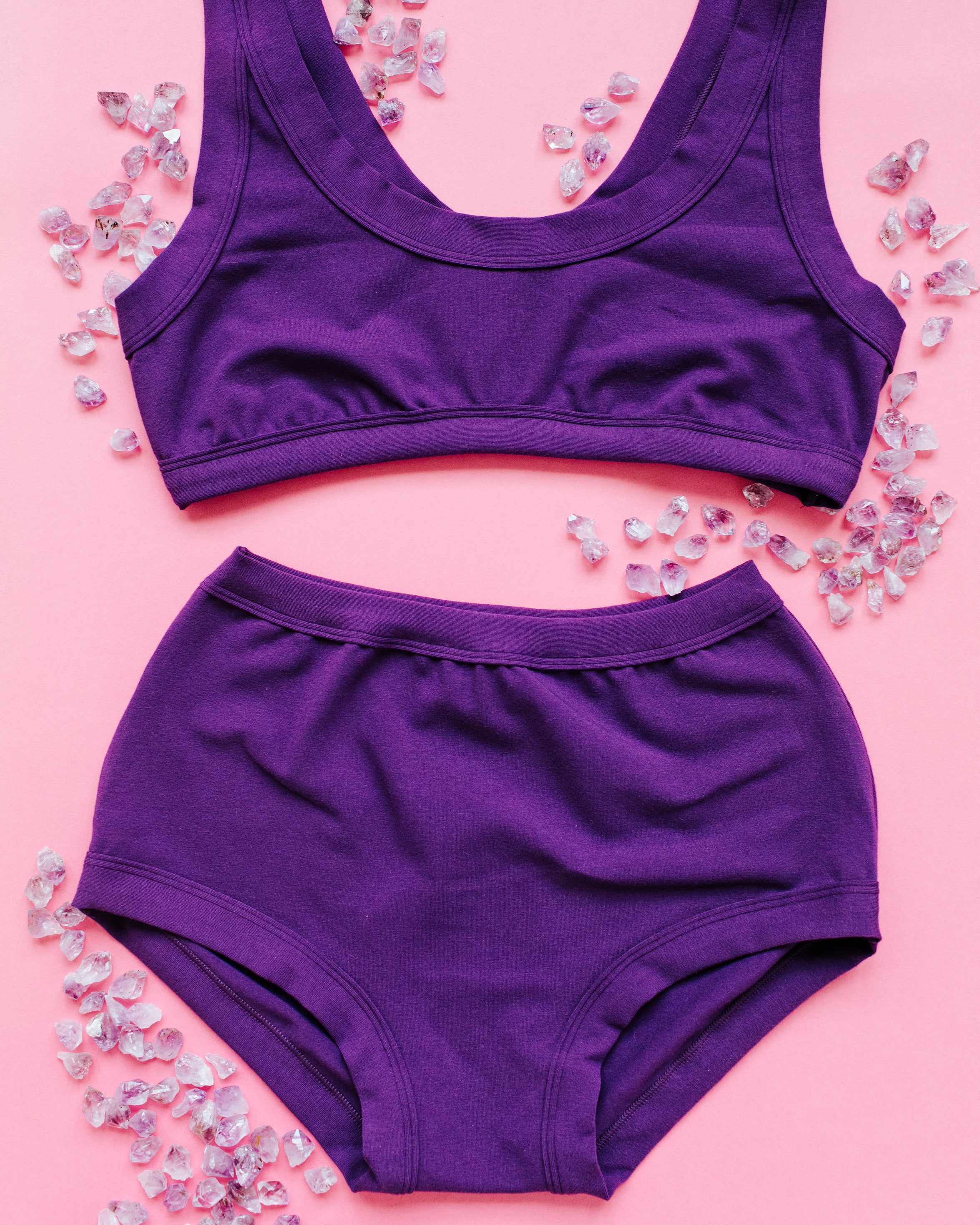 Flat lay of purple Deep Amethyst Original style underwear and Bralette on a pink surface with small amethyst stones surrounding.