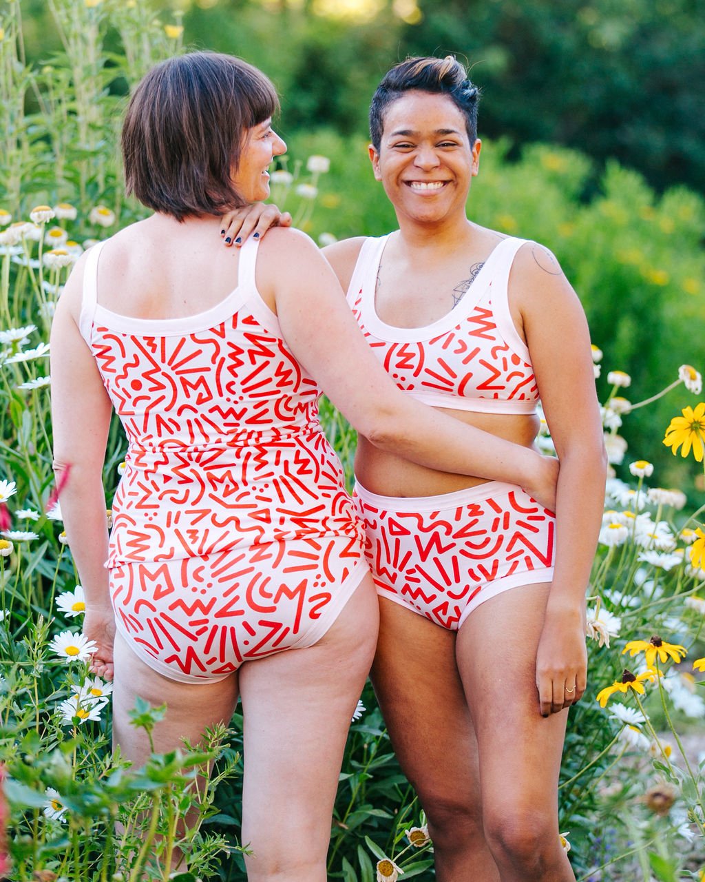 Two models smiling together wearing Thunderpants organic cotton Original style and Hipster style underwear, Camisole and Bralette in our Energy Vibes print: pink with large red squiggles.