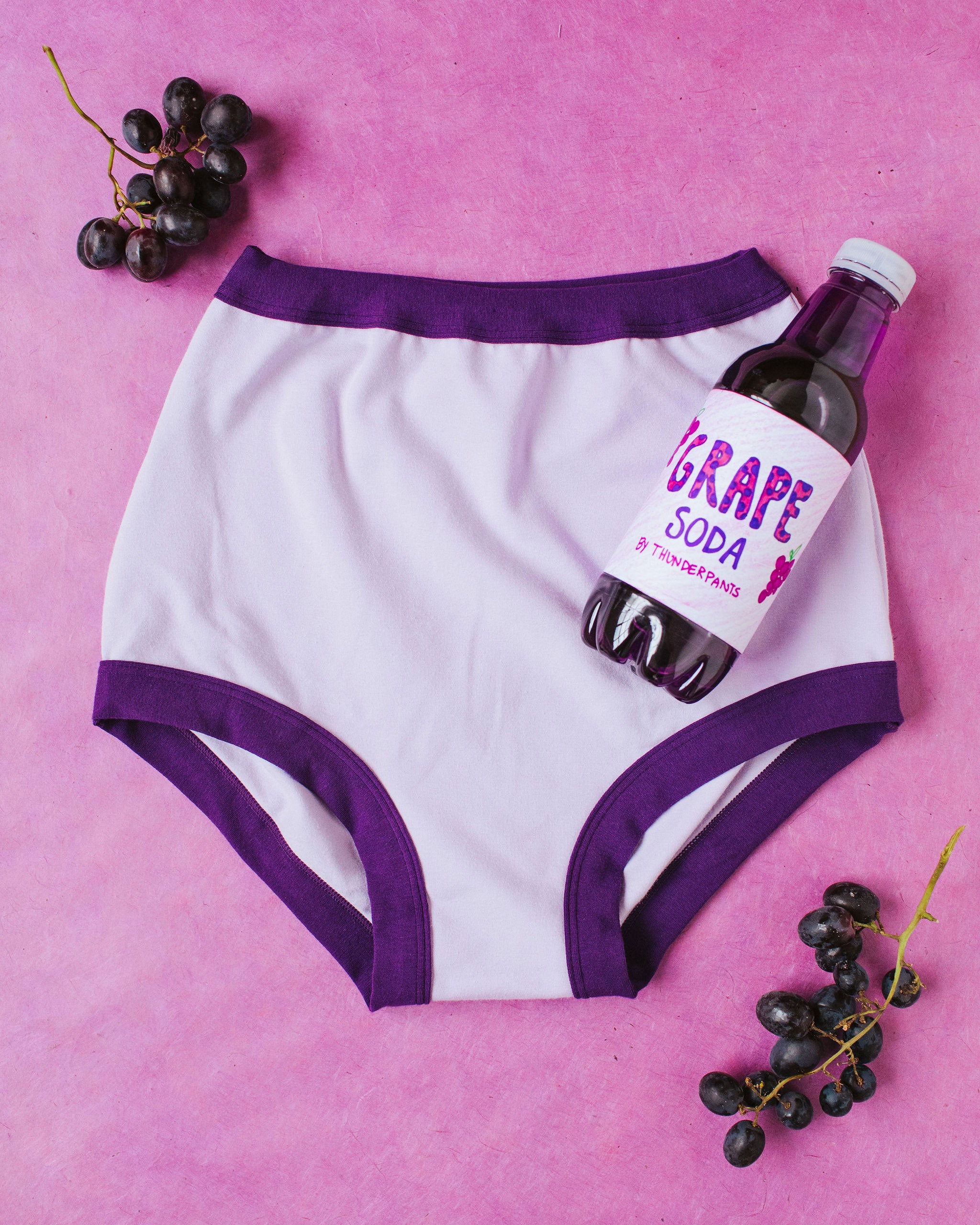 Flat lay with grape soda bottle on a purple surface of Sky Rise style underwear in Grape Soda: lavender color with dark purple binding.