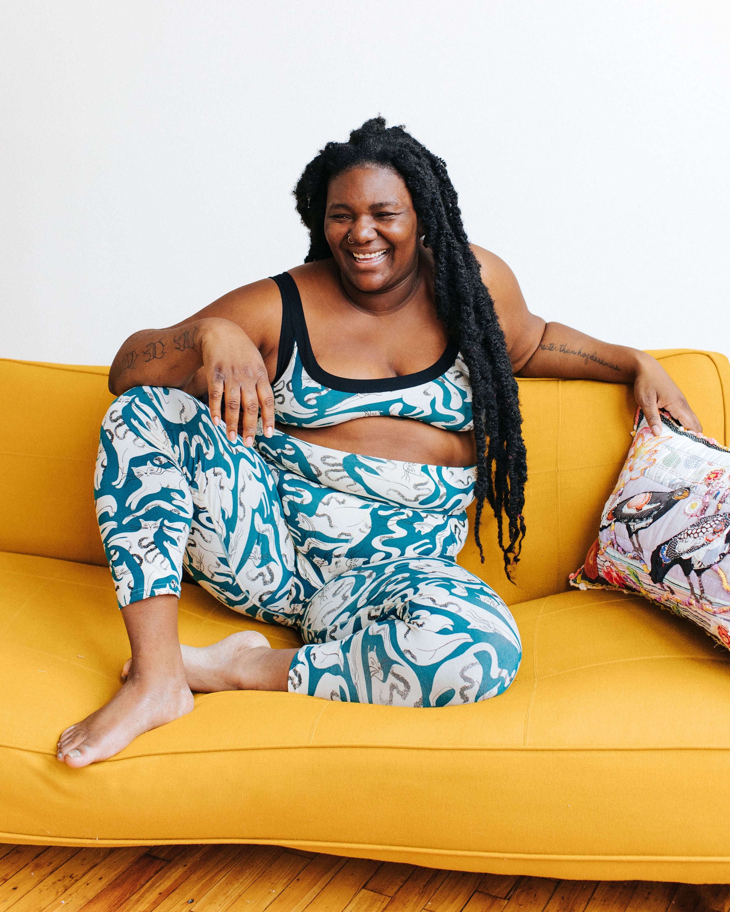 Model sitting on a yellow couch wearing Ankle Leggings and Bralette in Hey Meow! print - green with white cats.