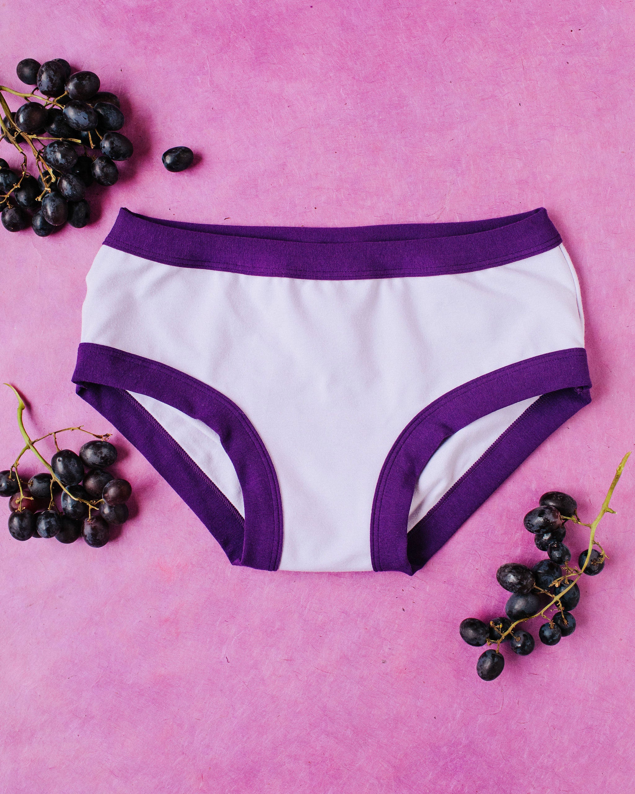 Flat lay with grapes on a purple surface of Grape Soda Hipsters style underwear: lavender color with dark purple binding.