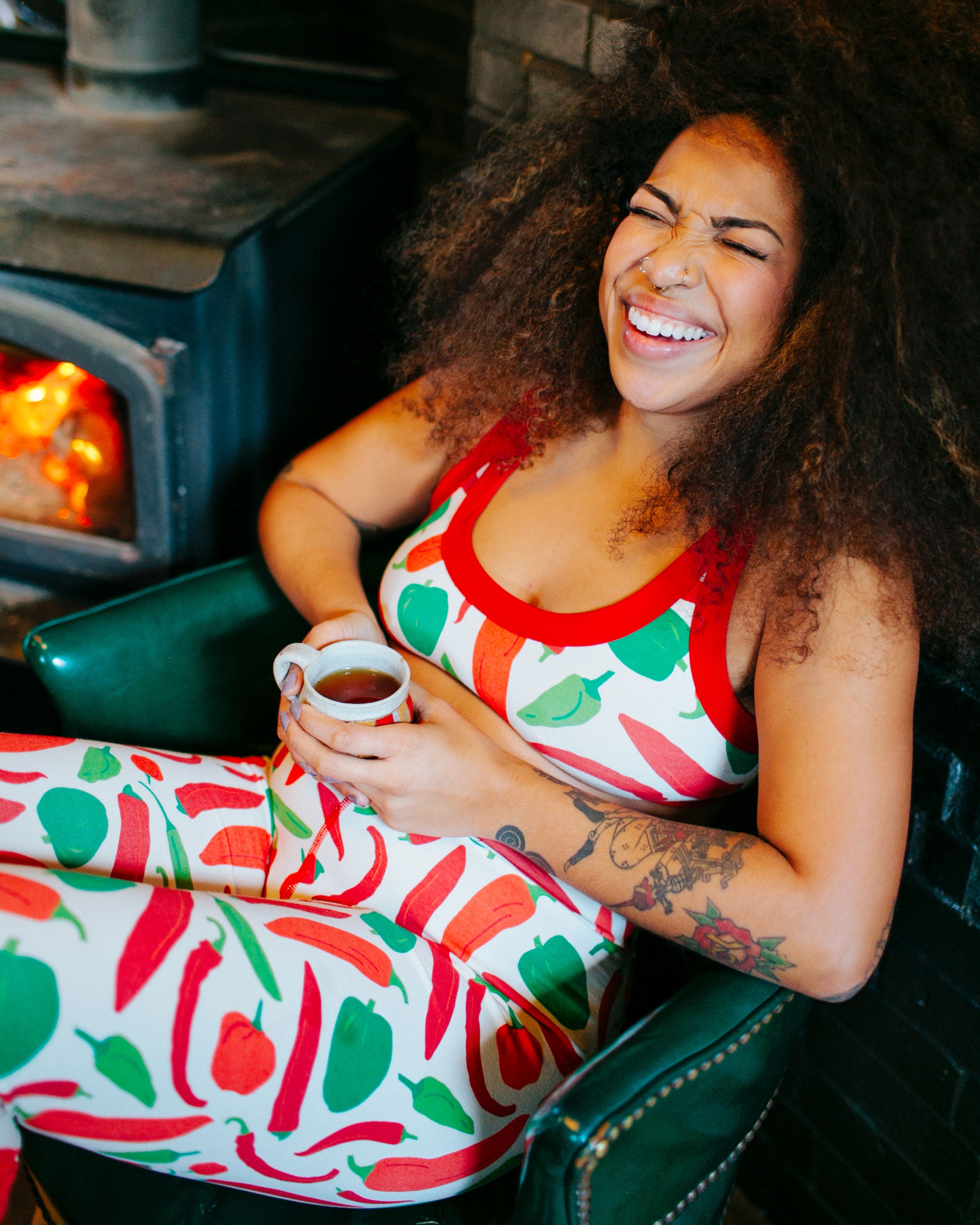 Model sitting by the fire laughing wearing Thunderpants organic cotton Bralette and Leggings in our Hot Pants print: various green, orange, and red peppers printed on Vanilla with red binding.