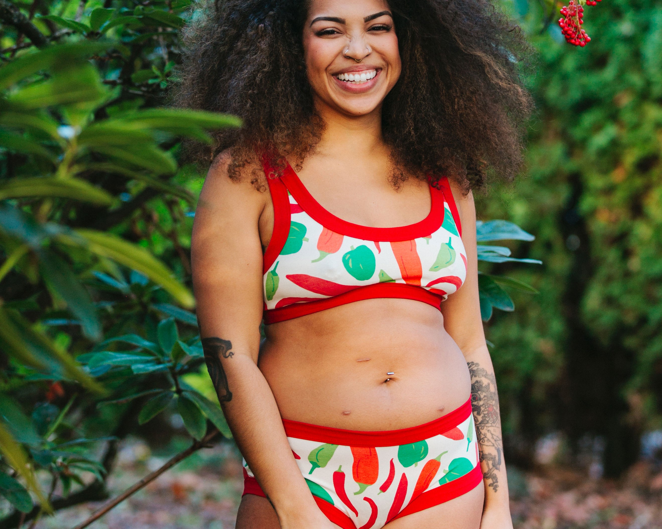 Model smiling big standing outside wearing Thunderpants organic cotton Hipster style underwear and Bralette in our Hot Pants print: various green, orange, and red peppers printed on Vanilla with red binding.