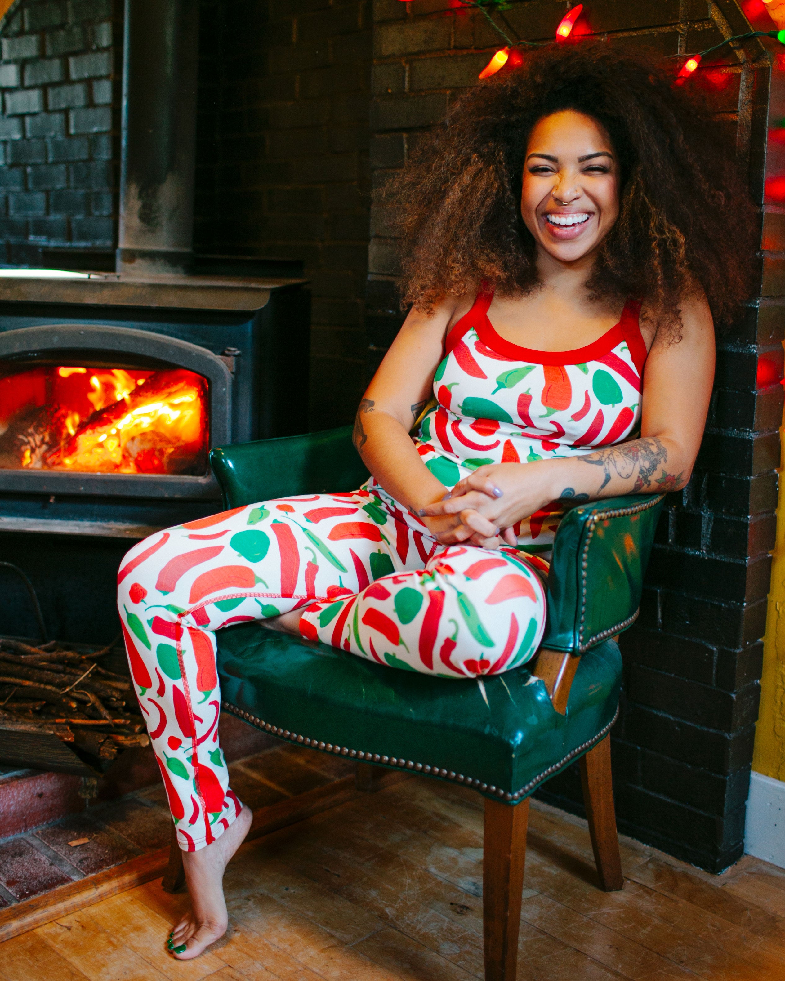 Model sitting by the fire and smiling into the camera wearing Thunderpants organic cotton Leggings and Camisole in our Hot Pants print: various green, orange, and red peppers printed on Vanilla with red binding.