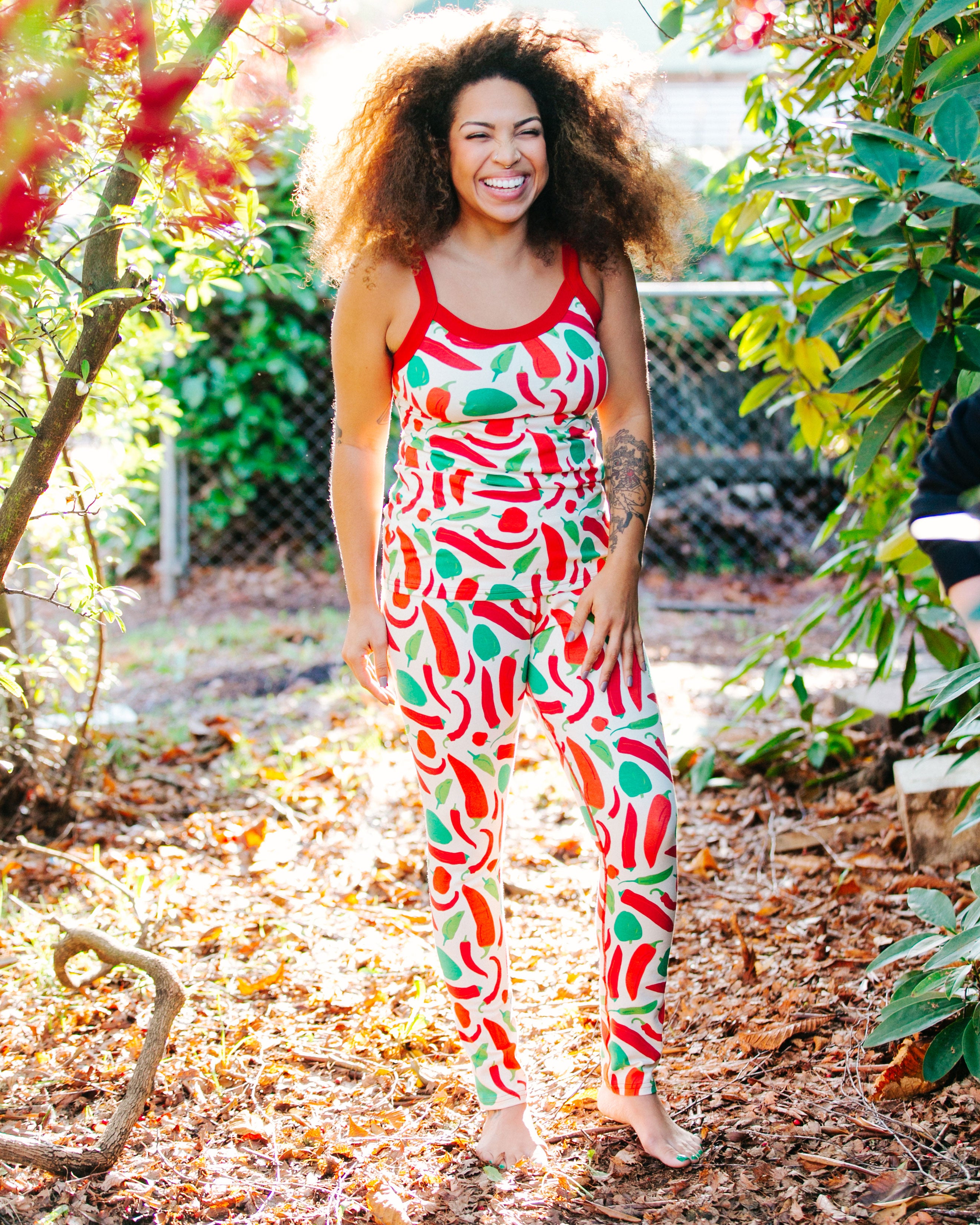 Model smiling big and standing outside wearing Thunderpants organic cotton Leggings and Camisole in our Hot Pants print: various green, orange, and red peppers printed on Vanilla with red binding.