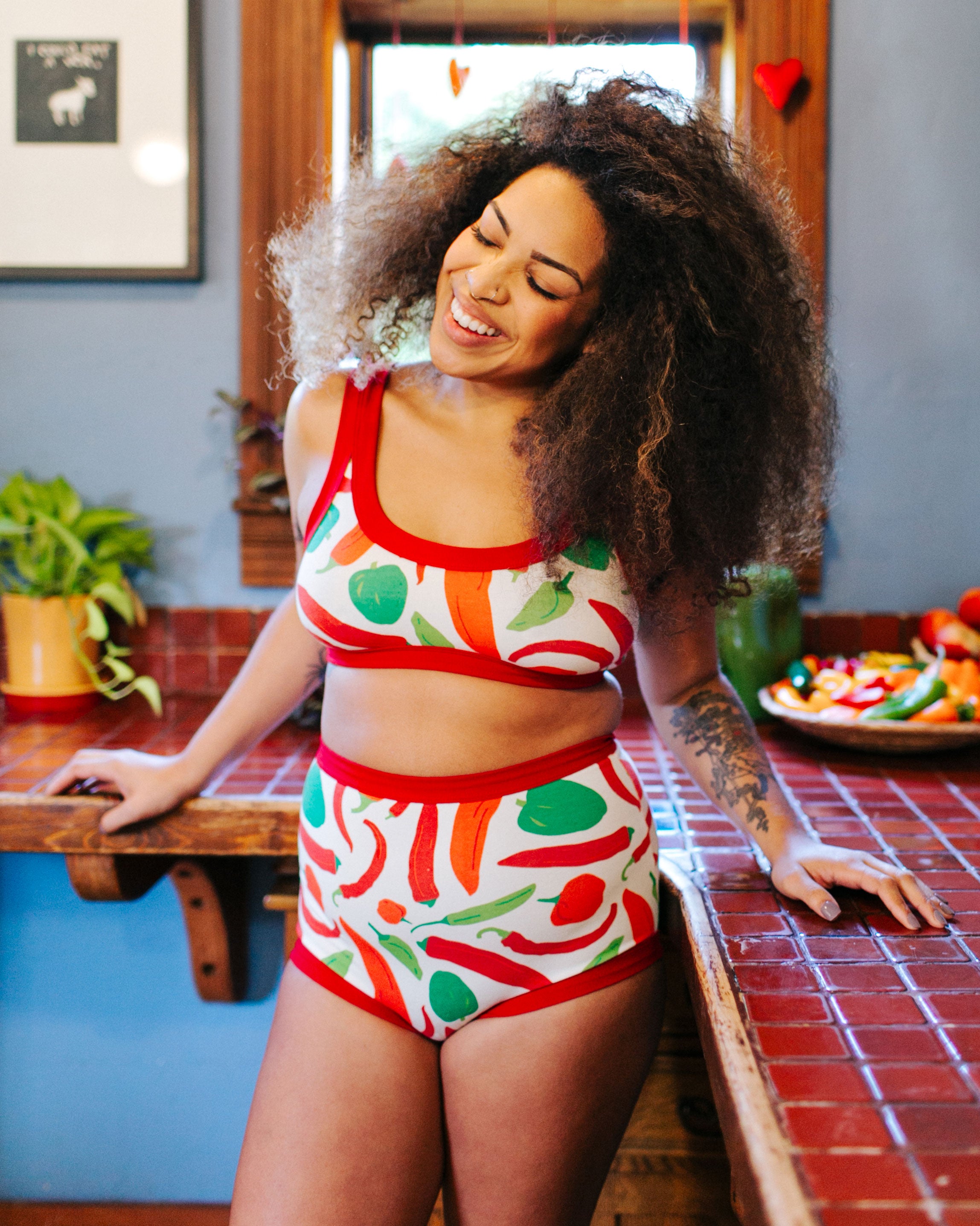 Model smiling standing in a kitchen wearing Thunderpants organic cotton Sky Rise style underwear and Bralette in our Hot Pants print: various green, orange, and red peppers printed on Vanilla with red binding.