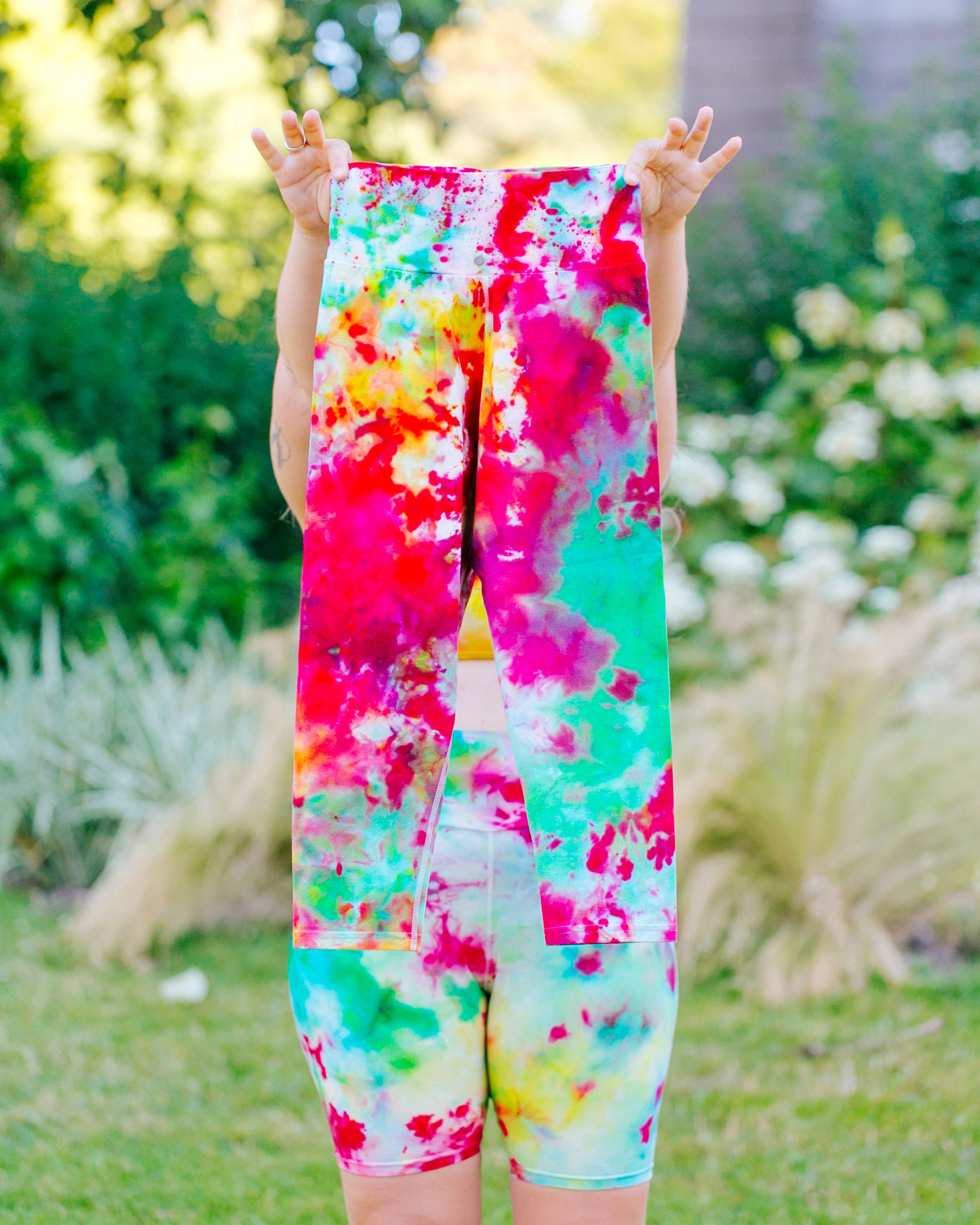 Model wearing Ice Dyed Bike Shorts holding up Ice Dyed 3/4 Length Leggings in a mix of pink, yellow, and green colors.