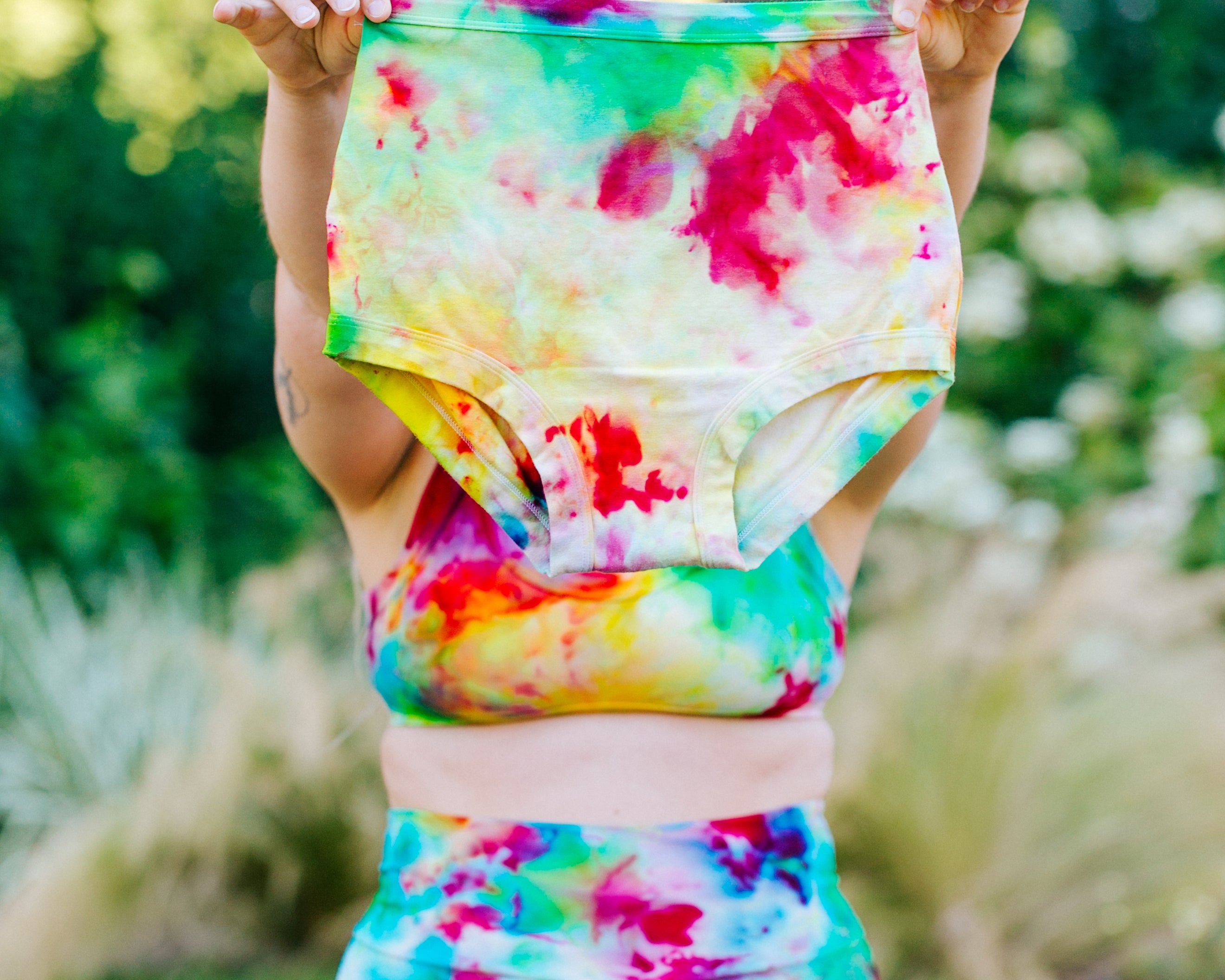 Model wearing Ice Dyed Bike Shorts and Bralette holding Ice Dyed Sky Rise style underwear in green, pink, and yellow colors.