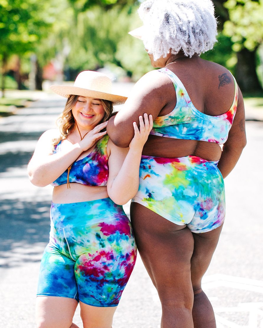 Two models smiling together wearing Thunderpants organic cotton High Rise Bike Shorts, Bralettes, and Original style underwear in hand dye Ice Dye multicolored tie-dye.