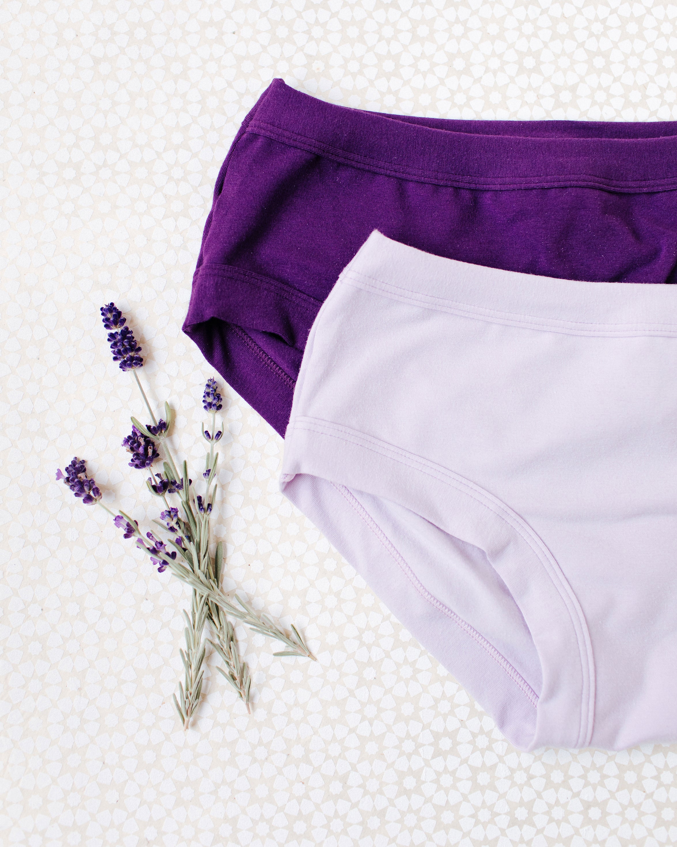 Flat lay of Light Lavender and Deep Amethyst Hipster style underwear on a white patterned surface with lavender sprigs.