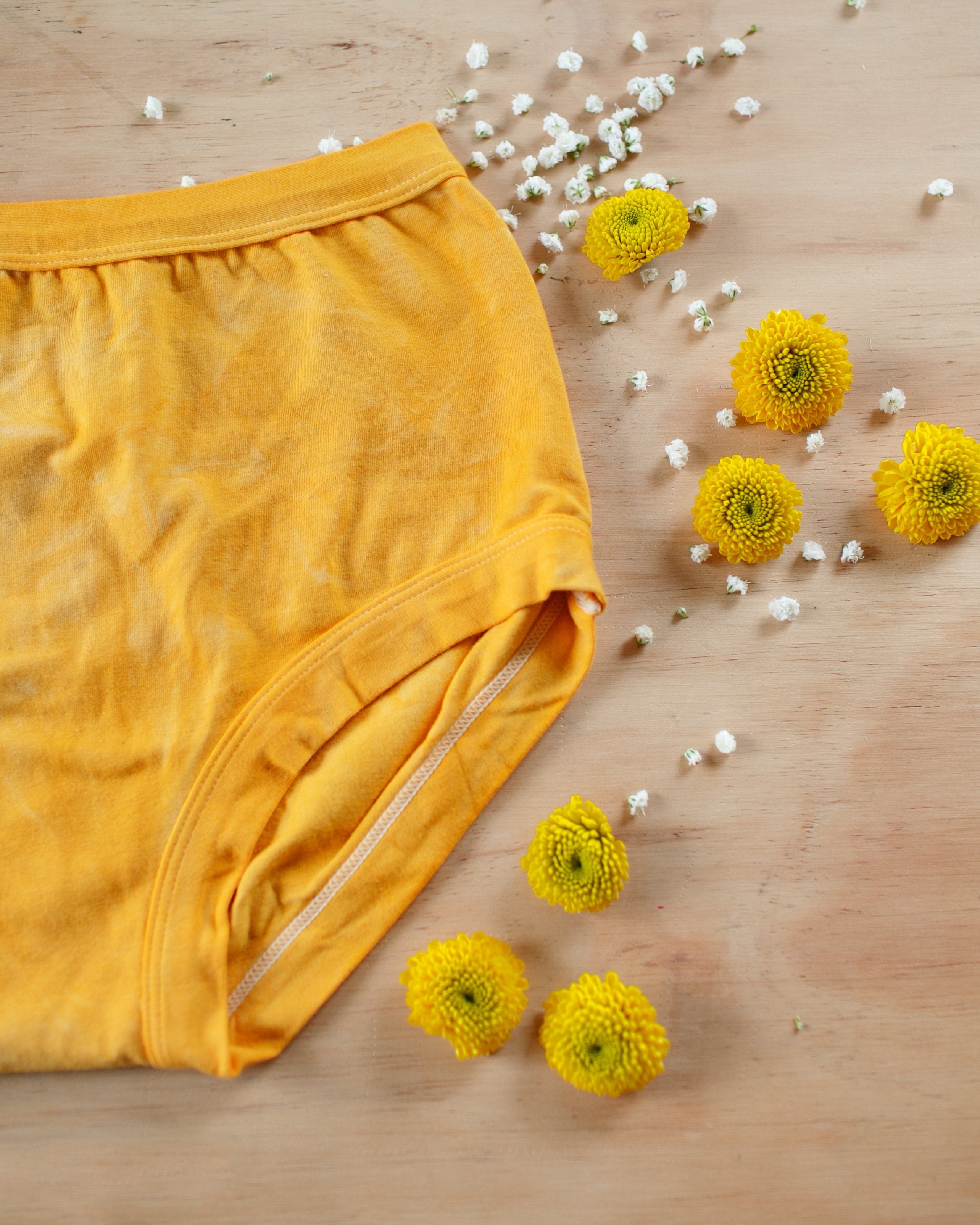 Flat-lay of Thunderpants organic cotton Original style underwear in limited edition hand dyed marigold color with marigolds and white flowers.