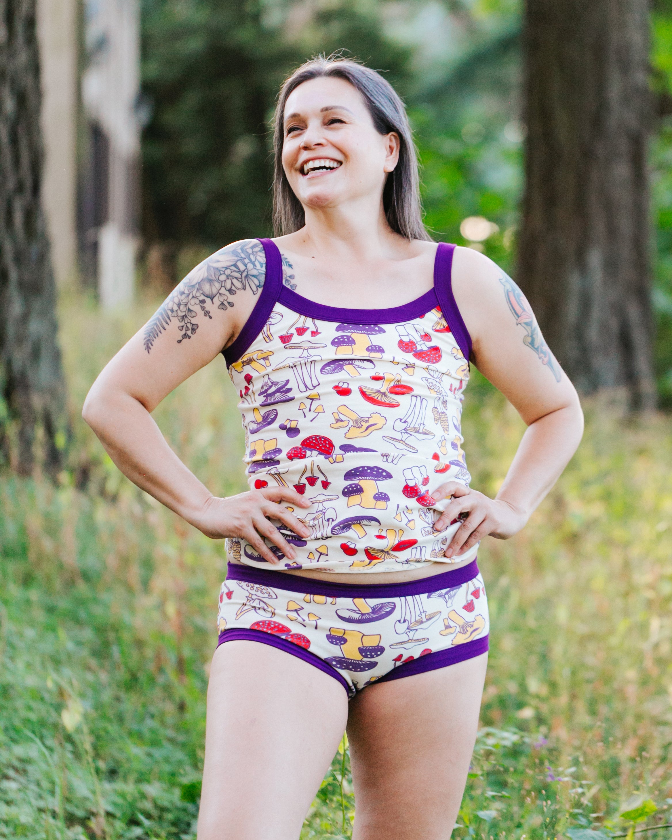 Model in the forest smiling and wearing a set of a Camisole and Hipster style underwear in Mushroom Magic: different kinds of mushrooms in red, yellow, and purple colors and dark purple binding.