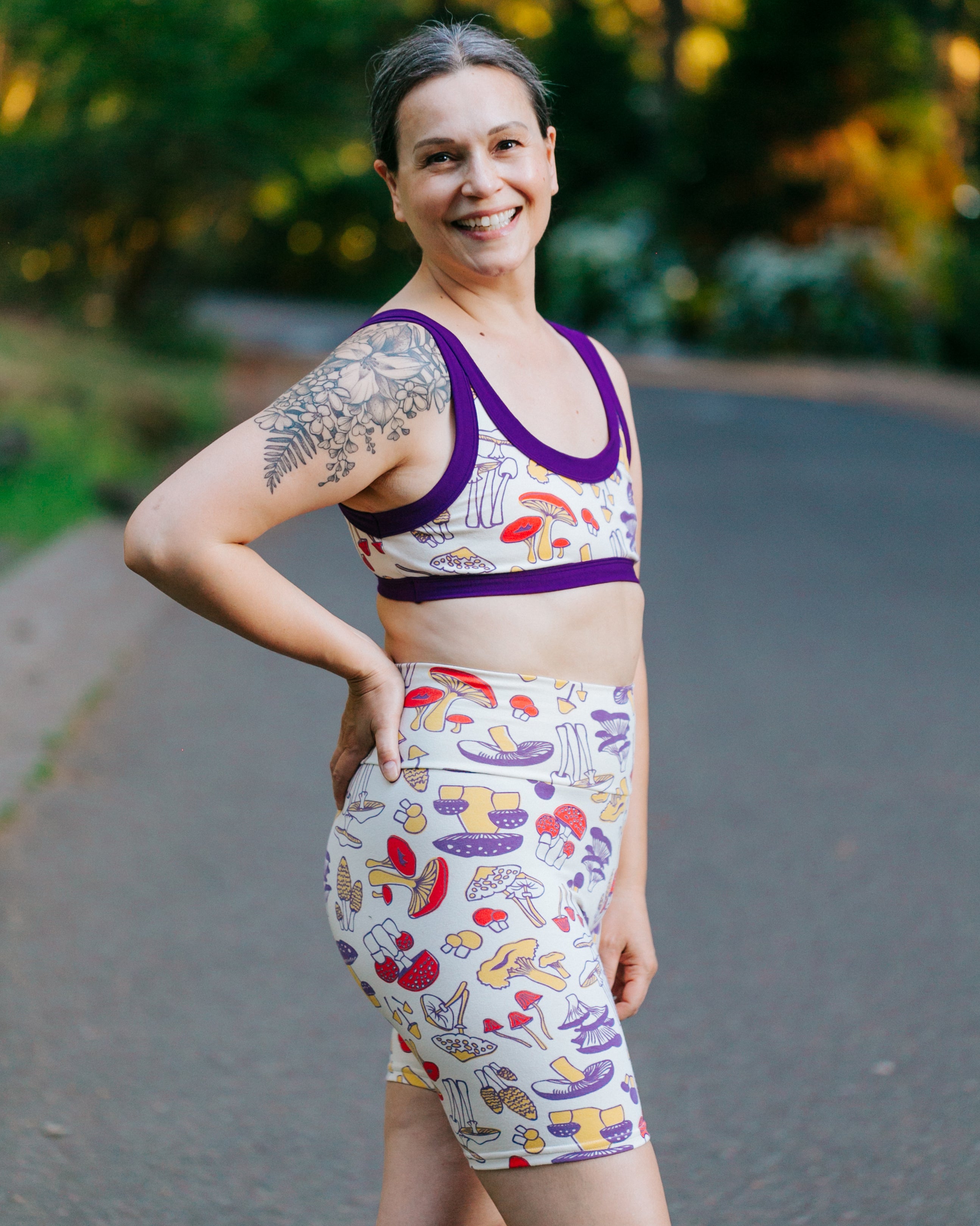 Model smiling wearing a set of Bike Shorts and Bralette in Mushroom Magic: different kinds of mushrooms in red, yellow, and purple colors.