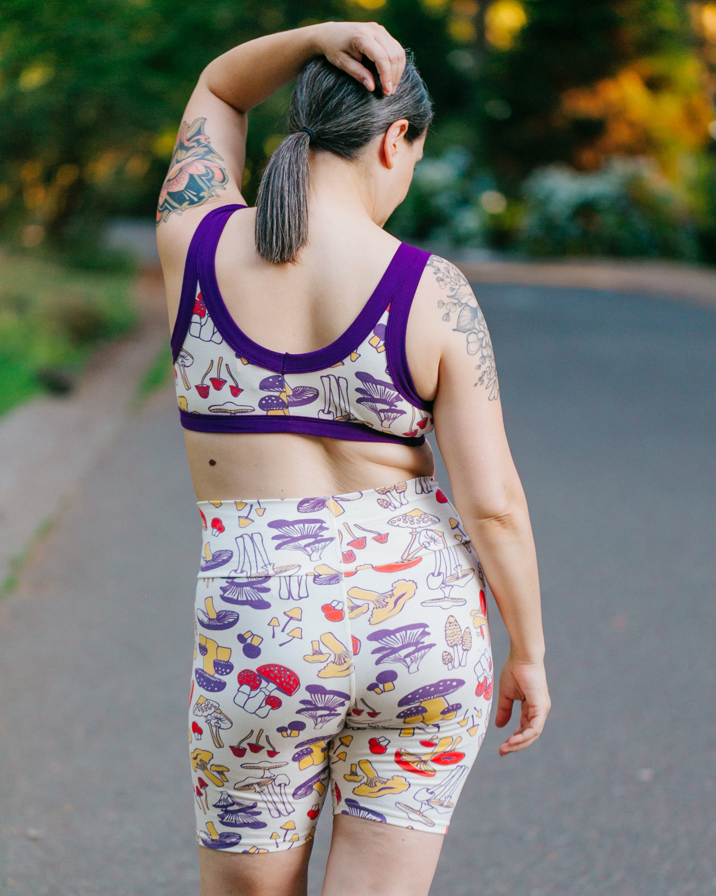 Model with her back to us wearing Bike Shorts and Bralette in Mushroom Magic print: different kinds of mushrooms in red, yellow, and purple colors.