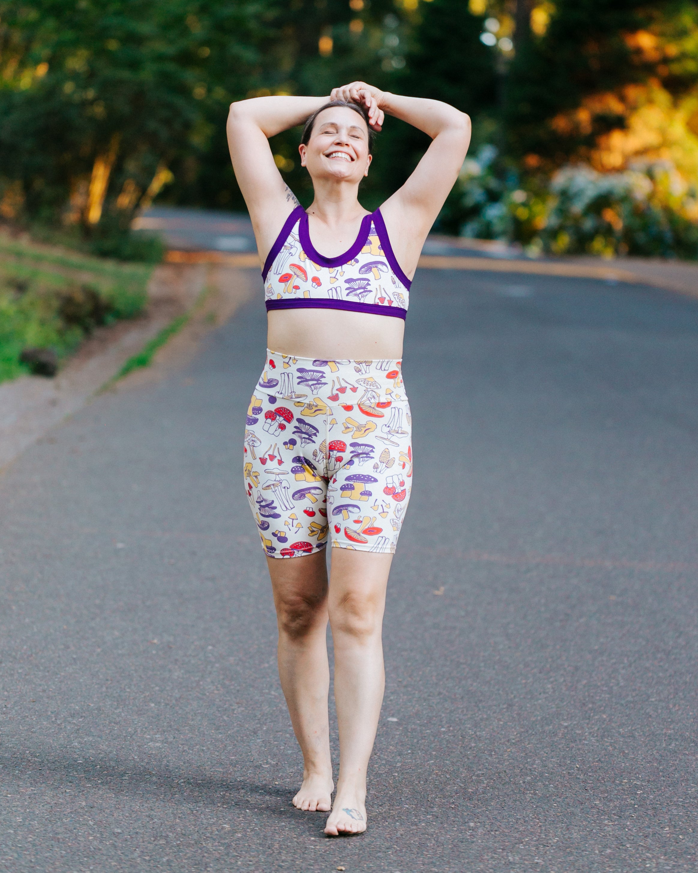 Model smiling big with her arms over her head wearing a set of Bike Shorts and Bralette in Mushroom Magic print: different kinds of mushrooms in yellow, red, and purple colors.
