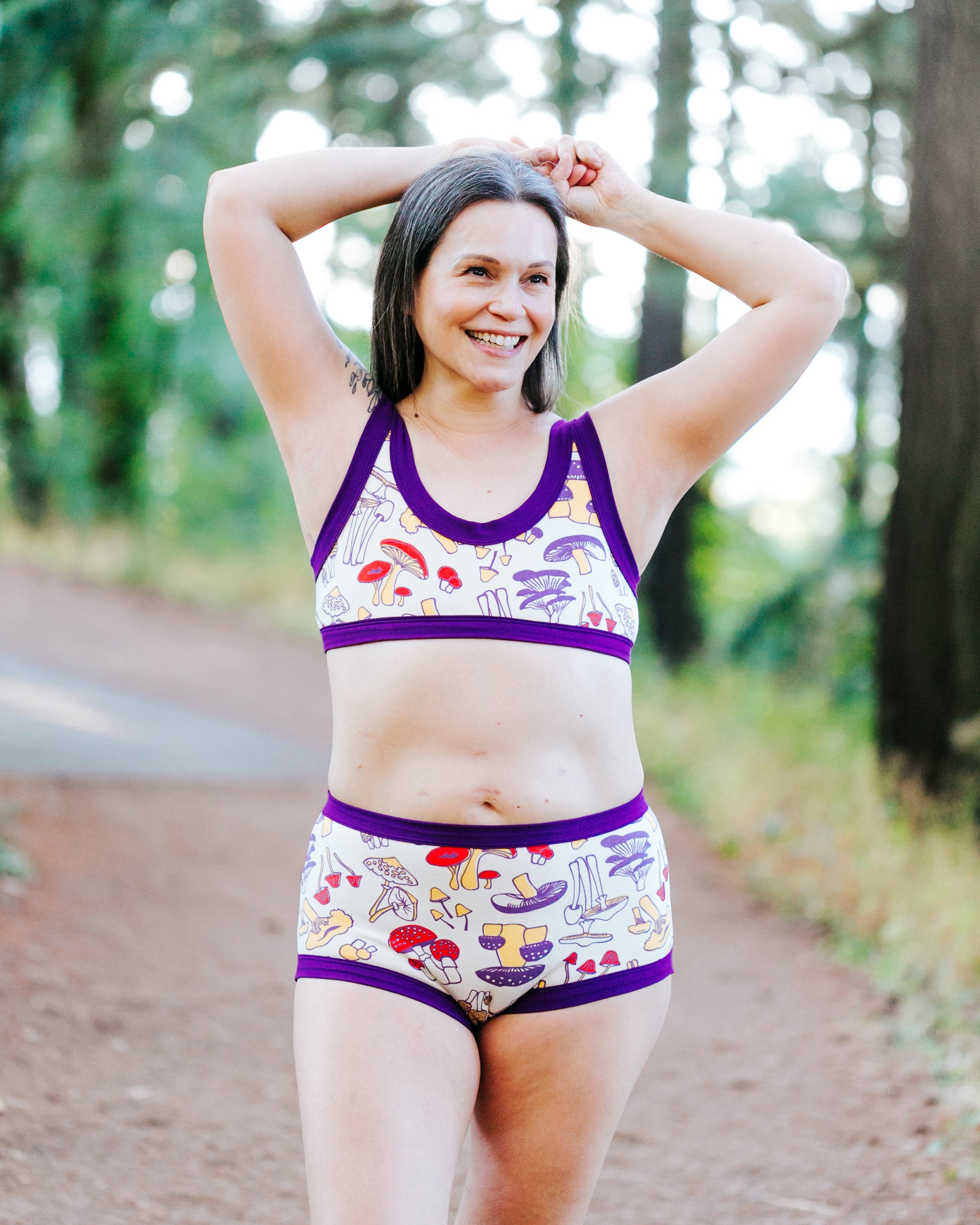 Model in the forest wearing a set of Bralette and Original style underwear in Mushroom Magic print: different kinds of mushrooms in red, yellow, and purple colors with dark purple binding.