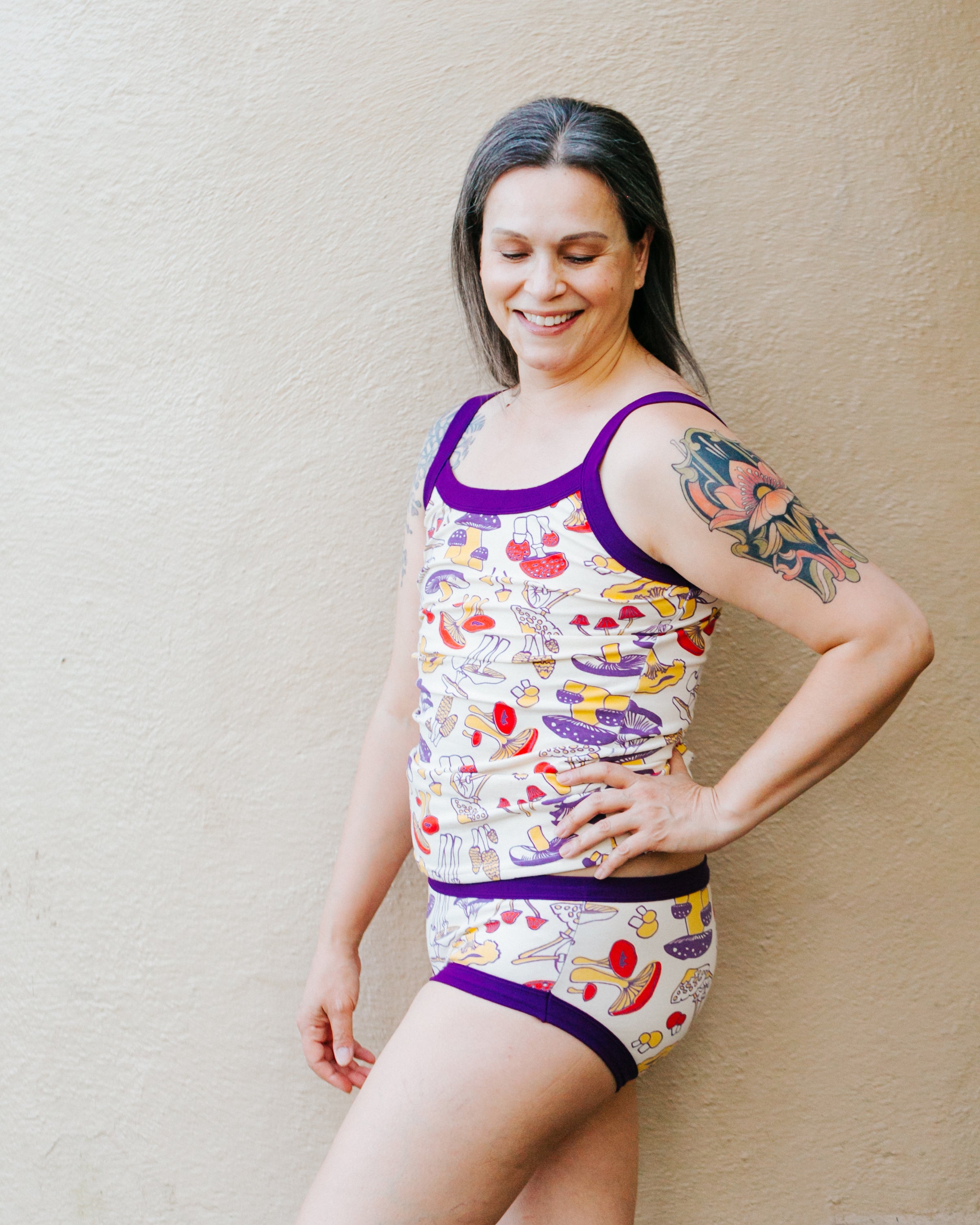 Model against a tan wall wearing a Camisole and Hipster style underwear in Mushroom Magic print: different kinds of mushrooms in red, yellow, and purple colors with dark purple binding.