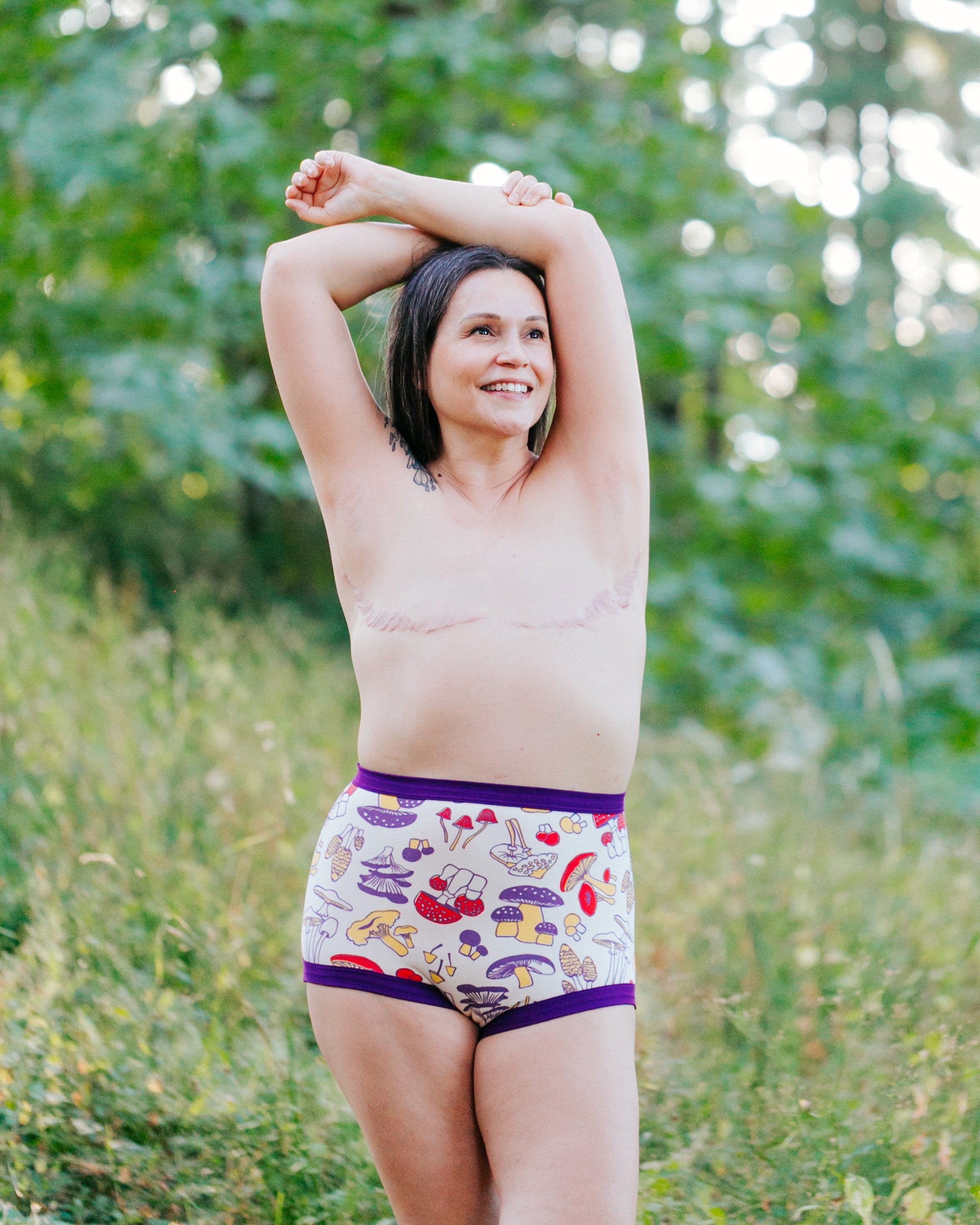 Model standing the forest with her arms over her head, smiling, proudly showing off her double vasectomy wearing Sky Rise style in Mushroom Magic print: different kinds of mushrooms in red, yellow, and purple colors with dark purple binding. 