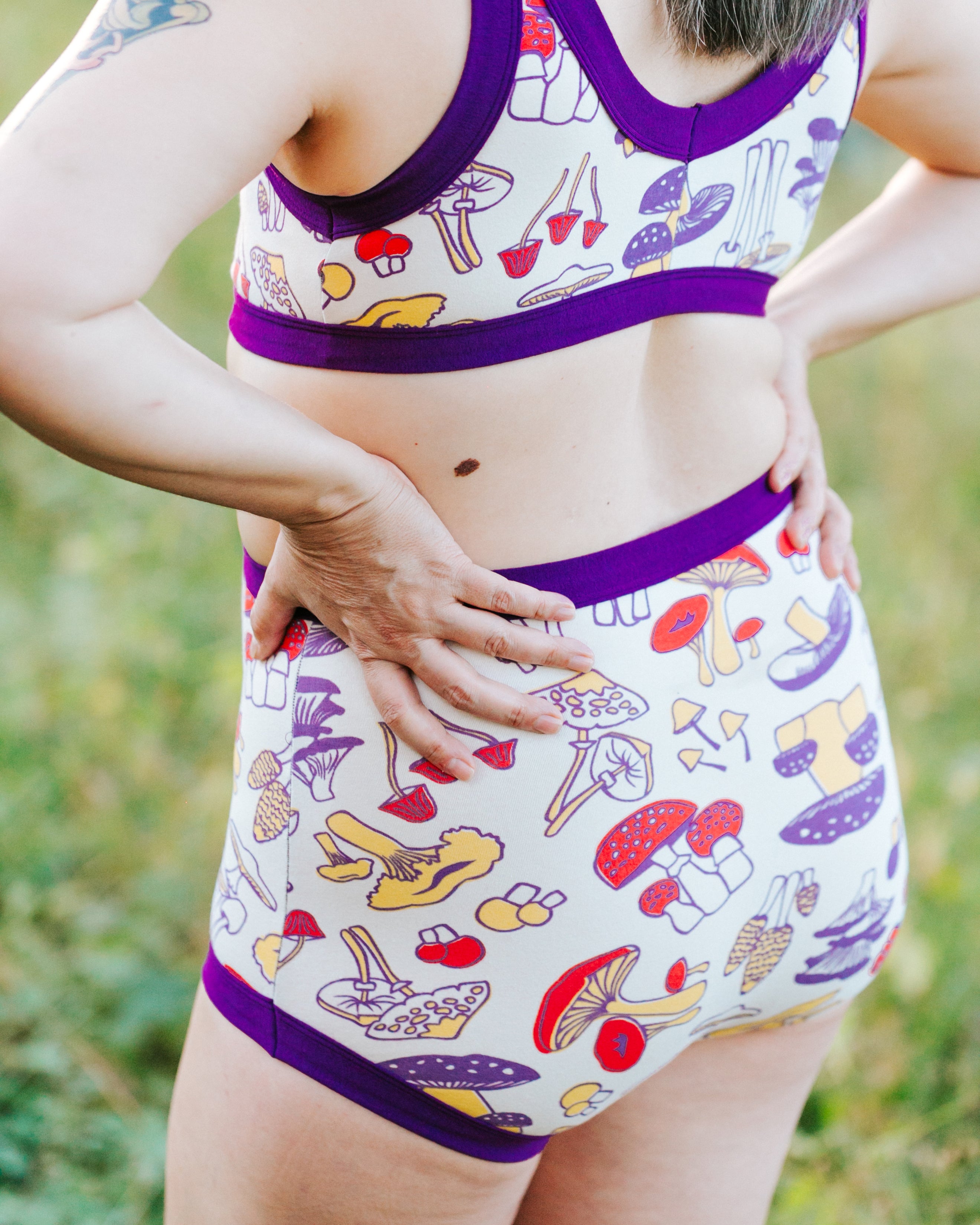 Close up of model's bum in Original style underwear in Mushroom Magic print: different kinds of mushrooms in red, yellow, and purple colors with dark purple binding.