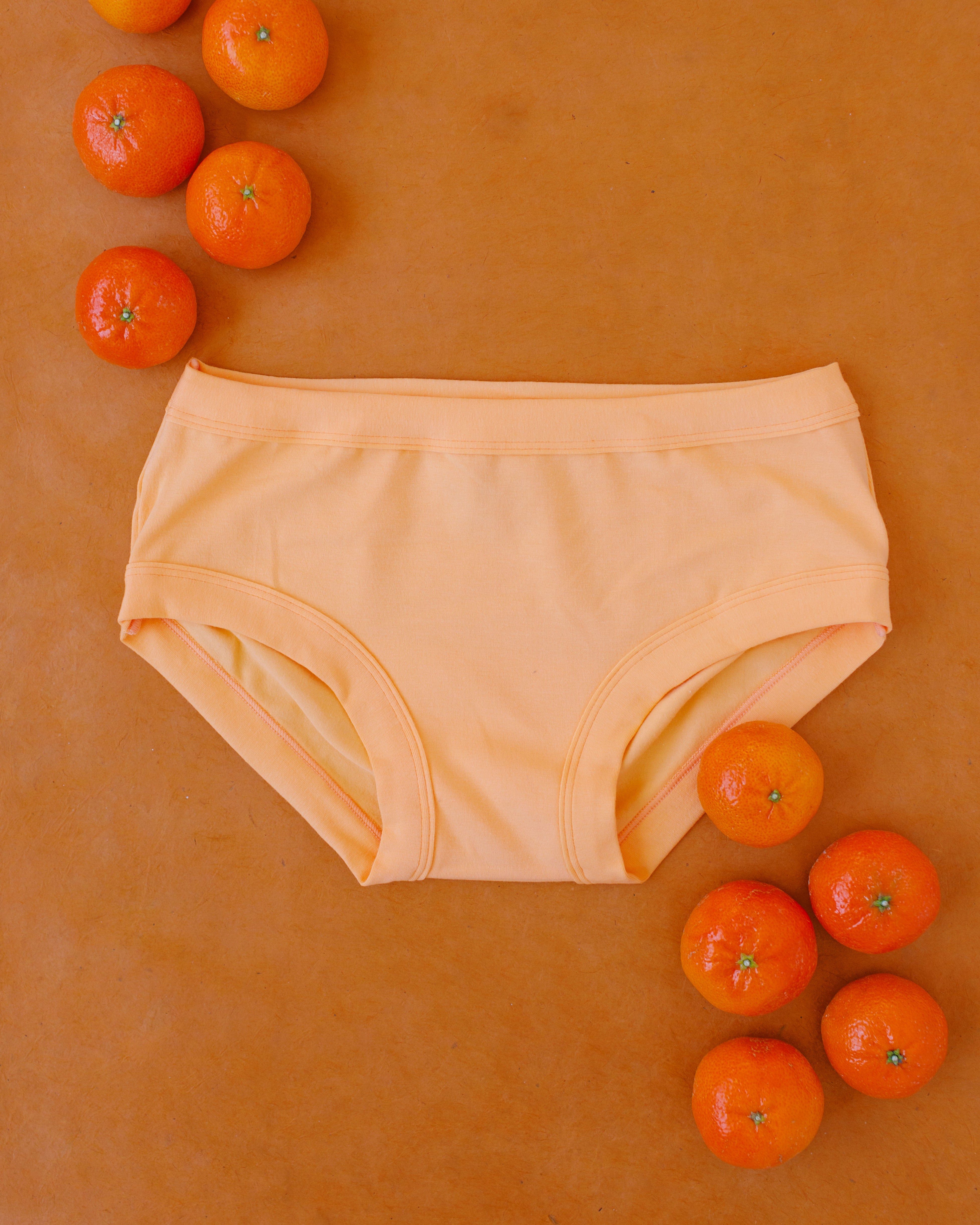 Flat lay of Orange Sherbet Hipster style underwear on an orange surface with oranges around it.