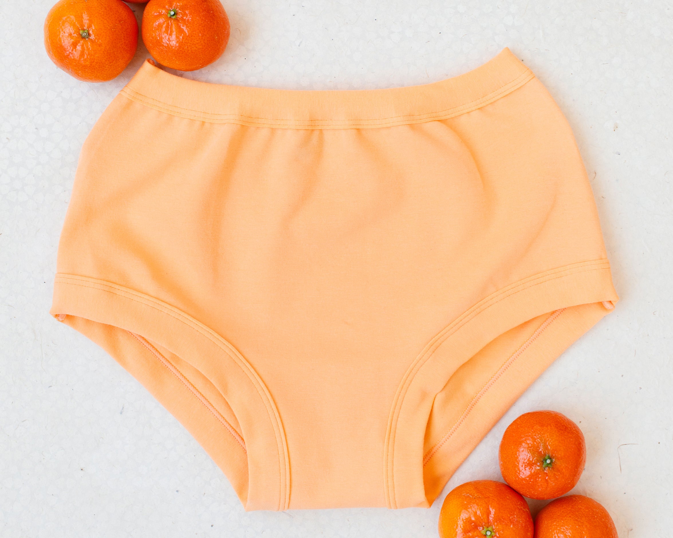Flat lay of Orange Sherbet Original style underwear on a white patterned surface with oranges around it.