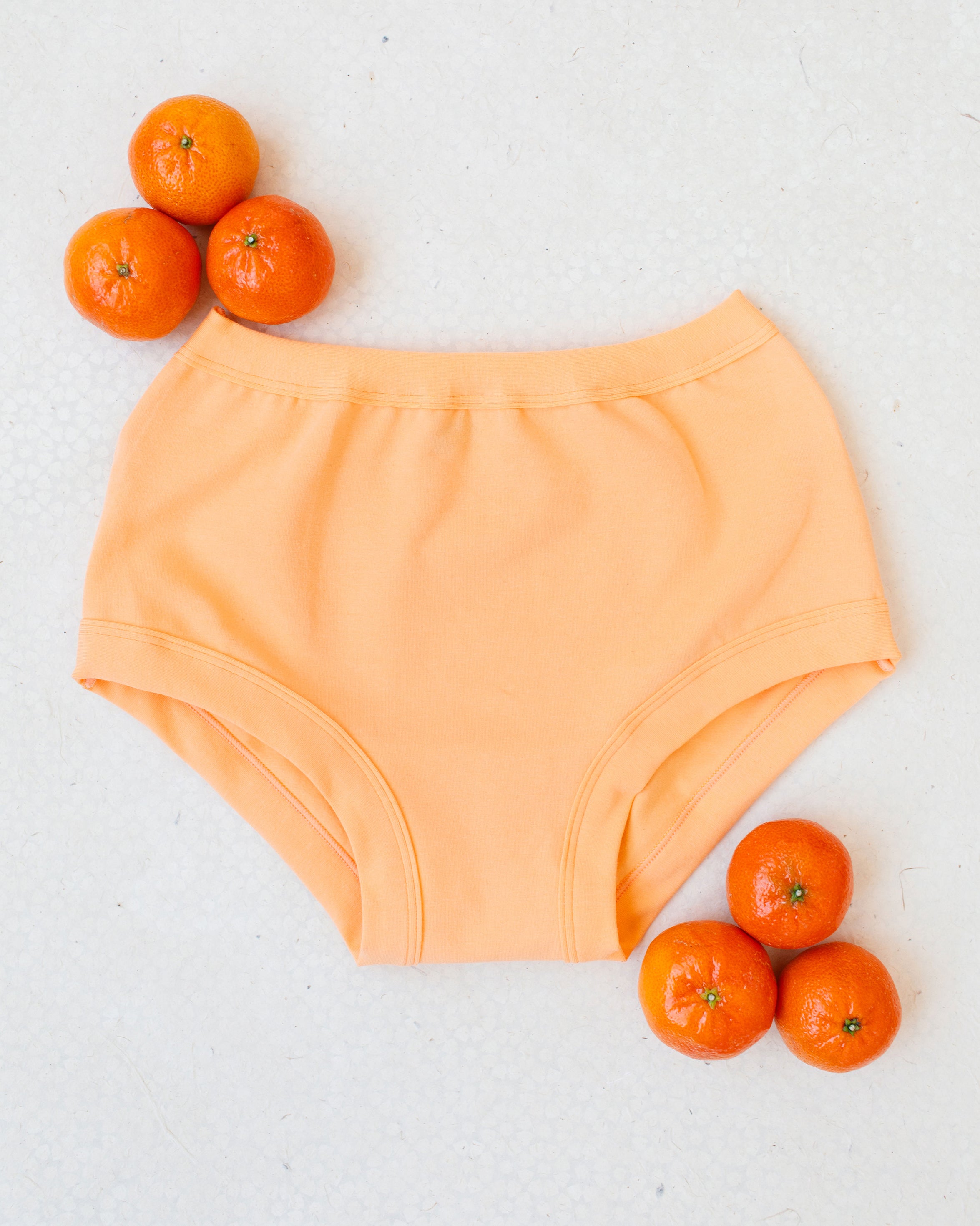 Flat lay of Orange Sherbet Original style underwear on a white patterned surface with oranges around it.
