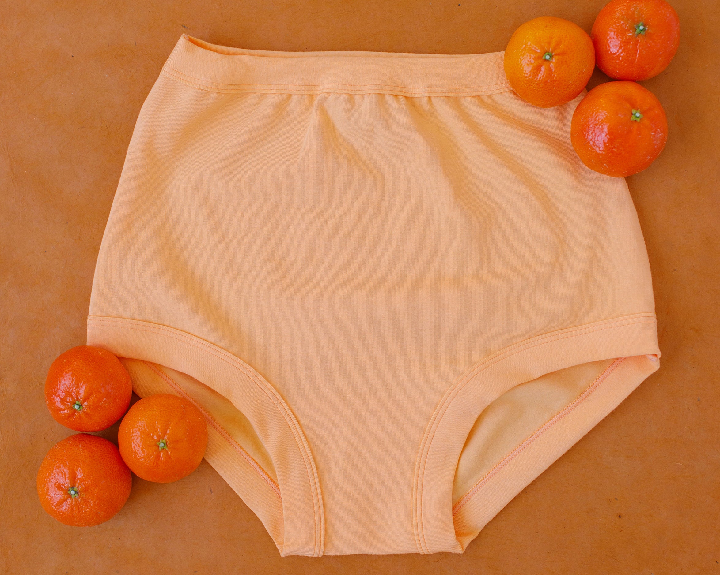 Flat lay of Orange Sherbet Sky Rise style underwear on a purple surface with oranges around it.