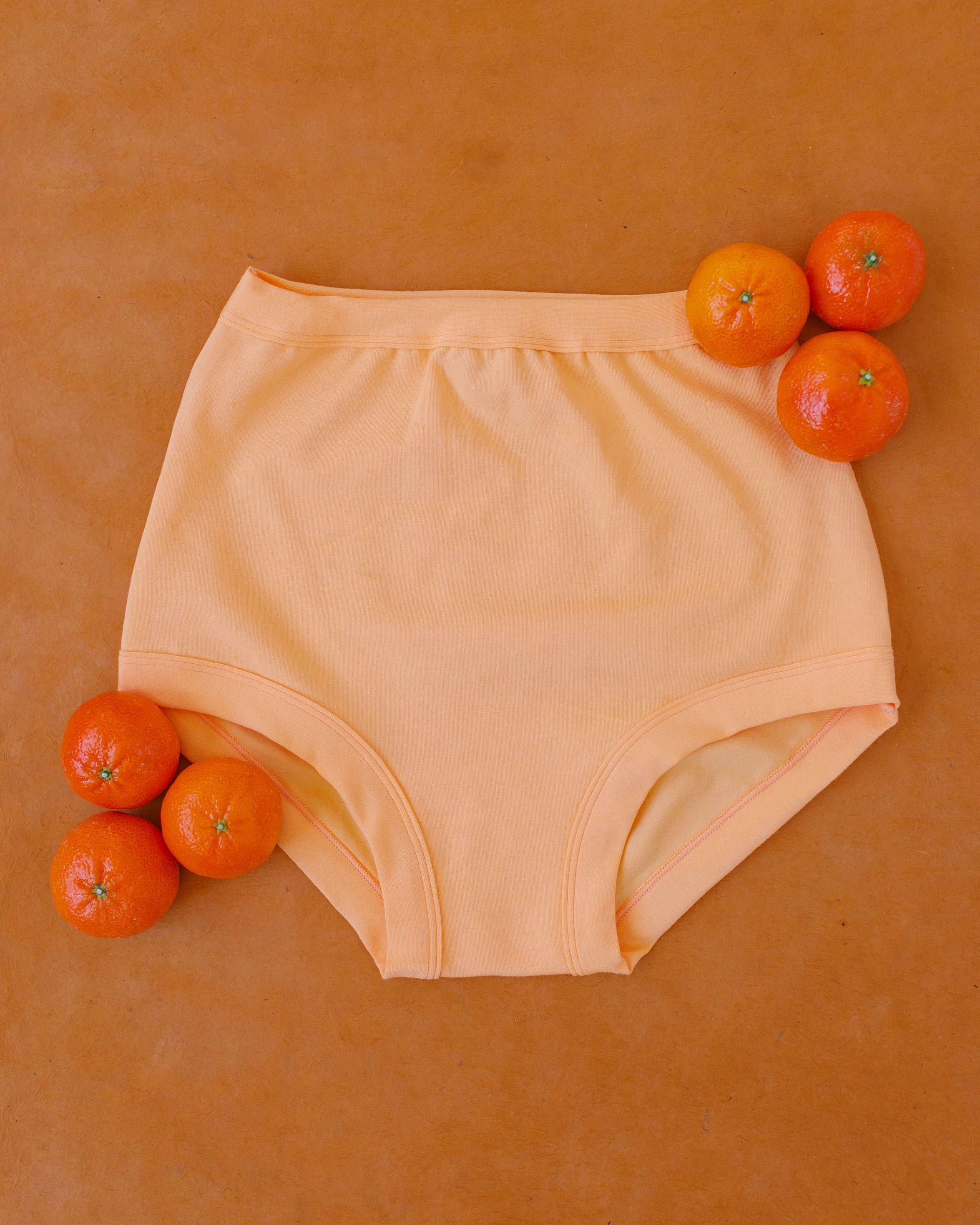 Flat lay of Orange Sherbet Sky Rise style underwear on a purple surface with oranges around it.
