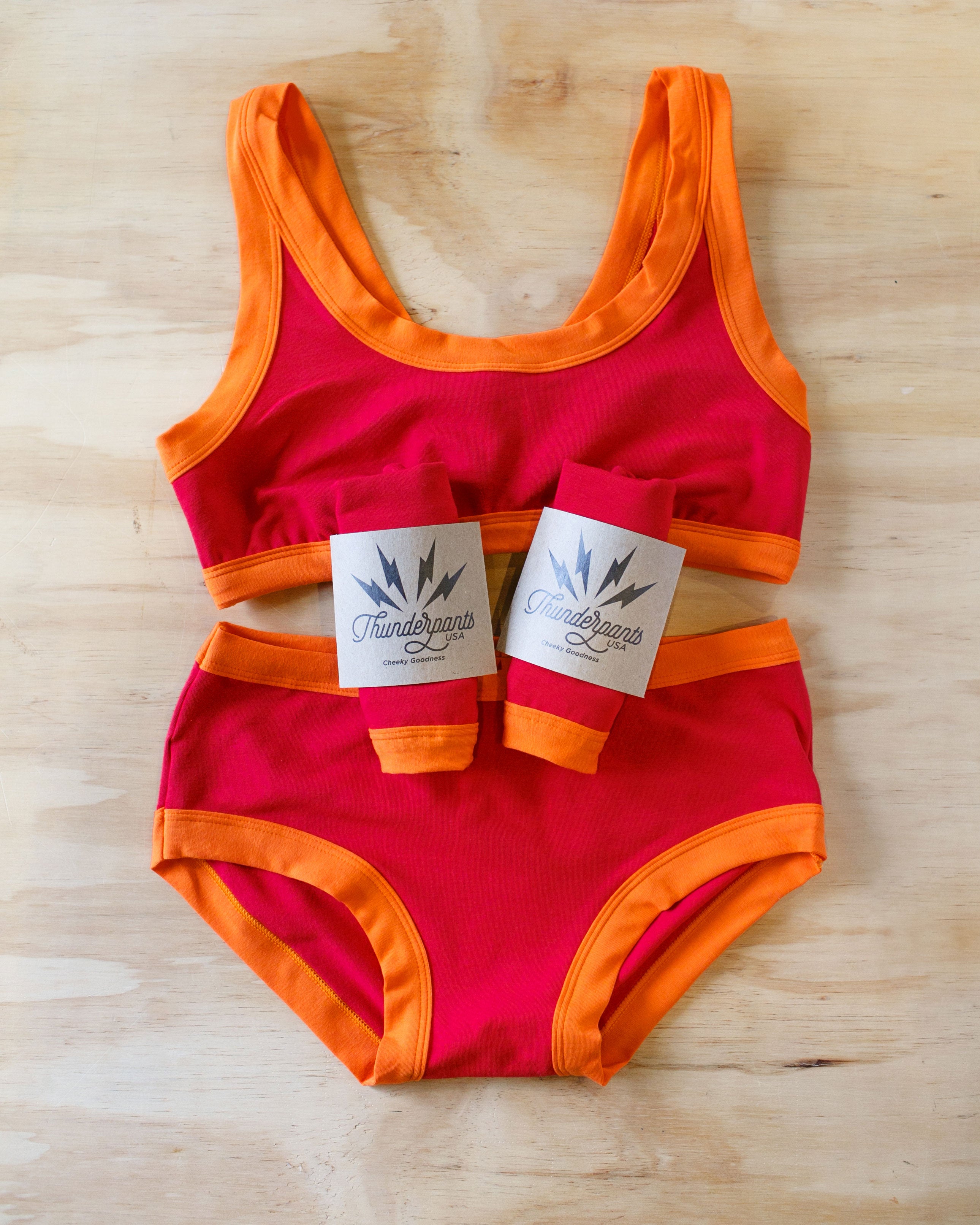 Flat lay on wood surface of Bralette, Hipster style underwear, and packaged underwear of Pants on Fire: red with orange bidning.