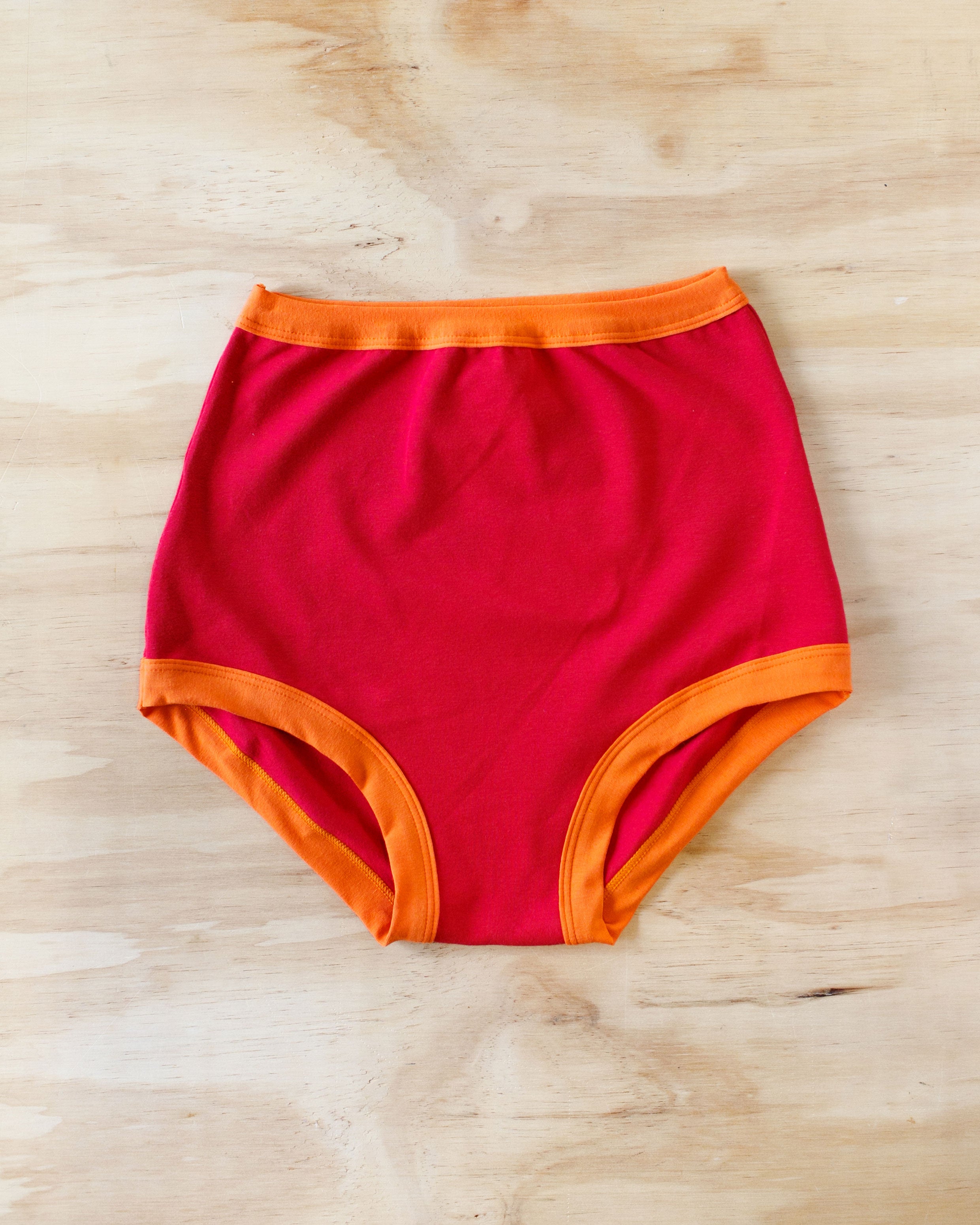 Flat lay on wood surface of Sky Rise style Pants on Fire: red with orange binding.
